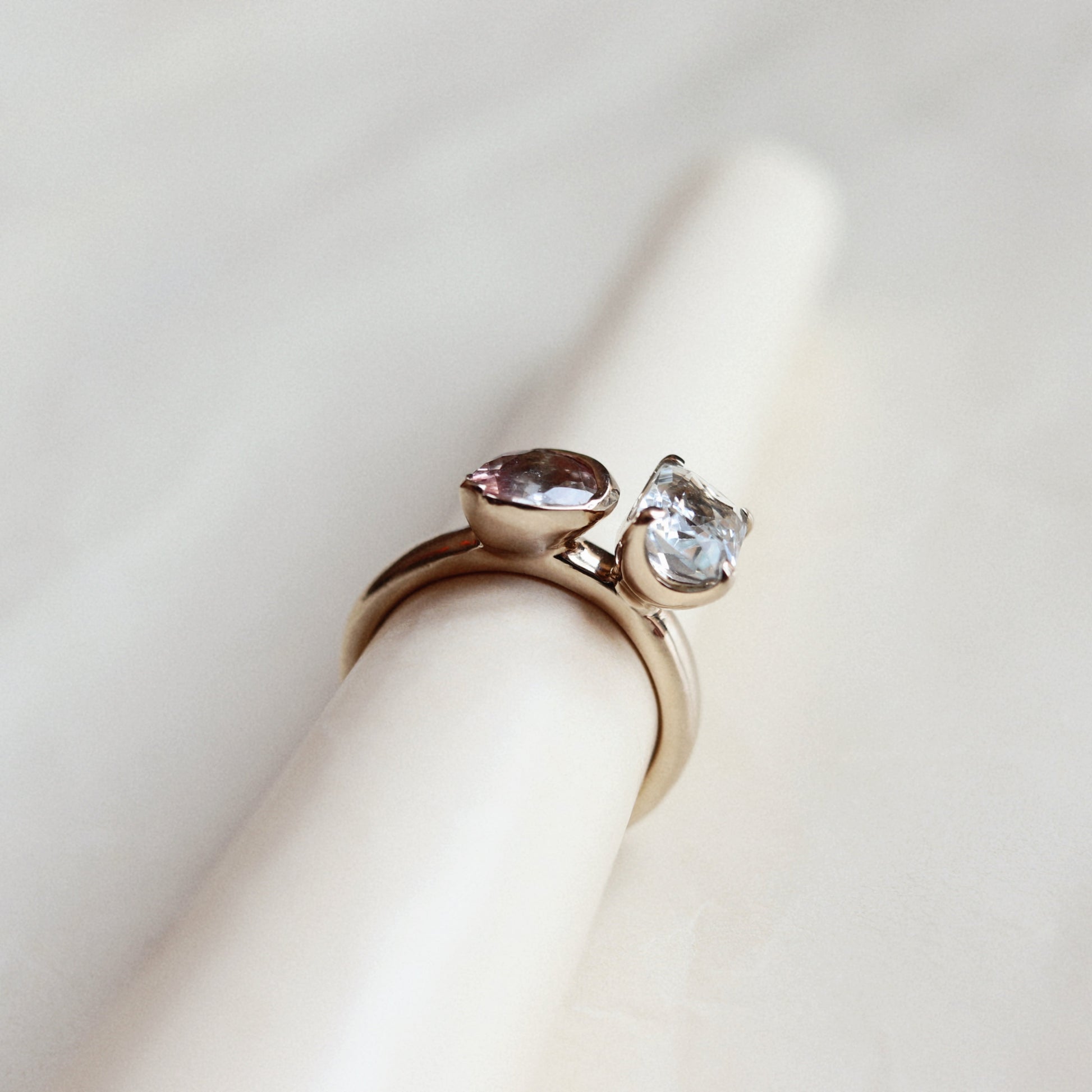A rose gold ring with two marquise-cut gemstones, one pinkish and one clear, displayed on a cream-colored ring stand against a soft, light background.