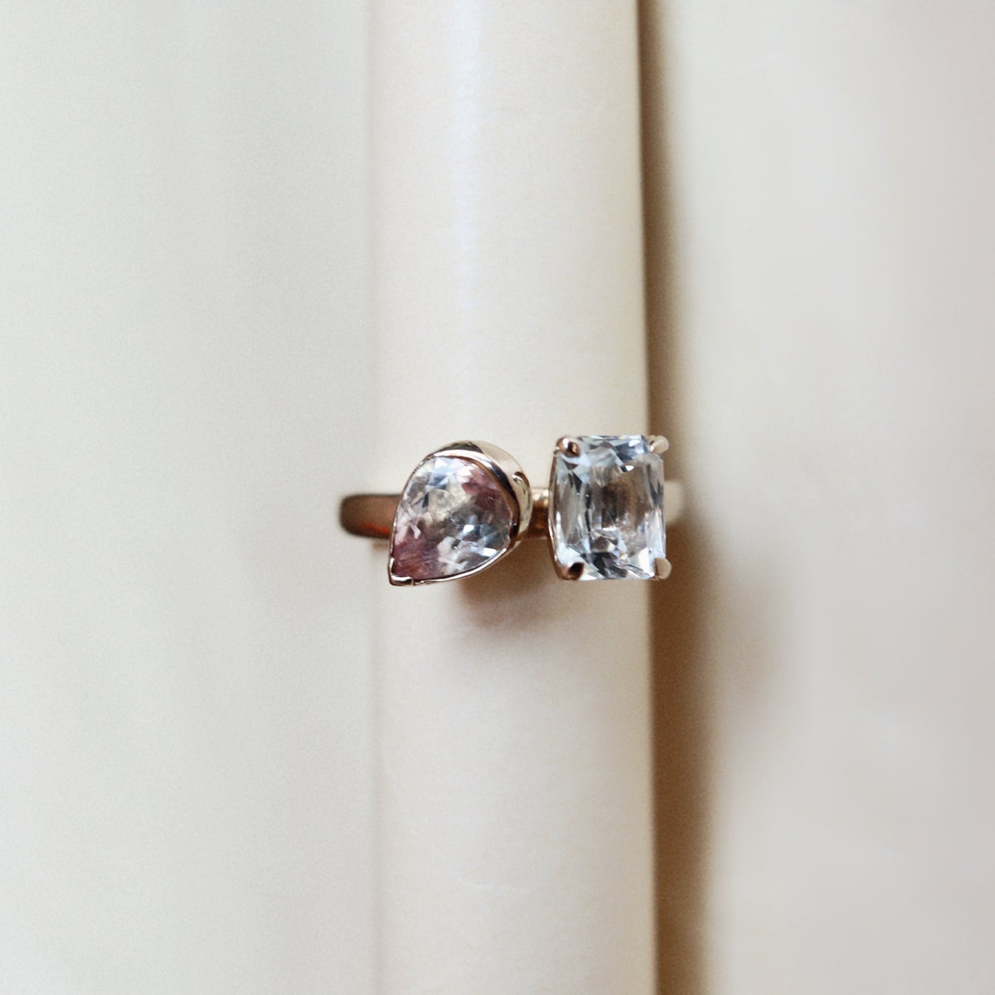 A pair of gold rings with clear gemstones, one pear-cut and one rectangular cushion-cut, displayed on a cream-colored ring stand.