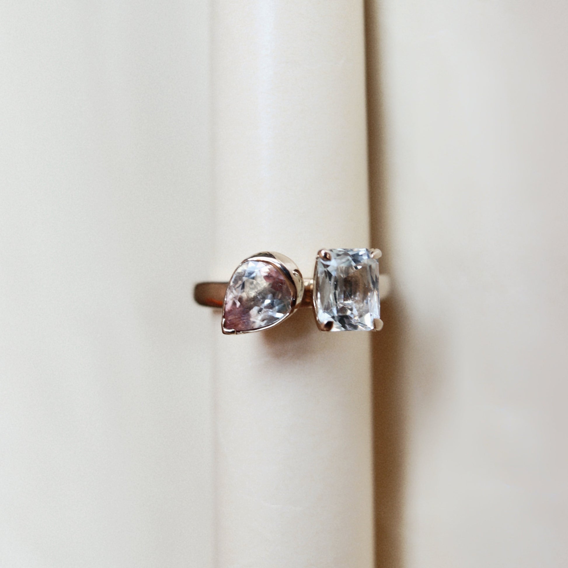 A pair of gold rings with clear gemstones, one pear-cut and one rectangular cushion-cut, displayed on a cream-colored ring stand.