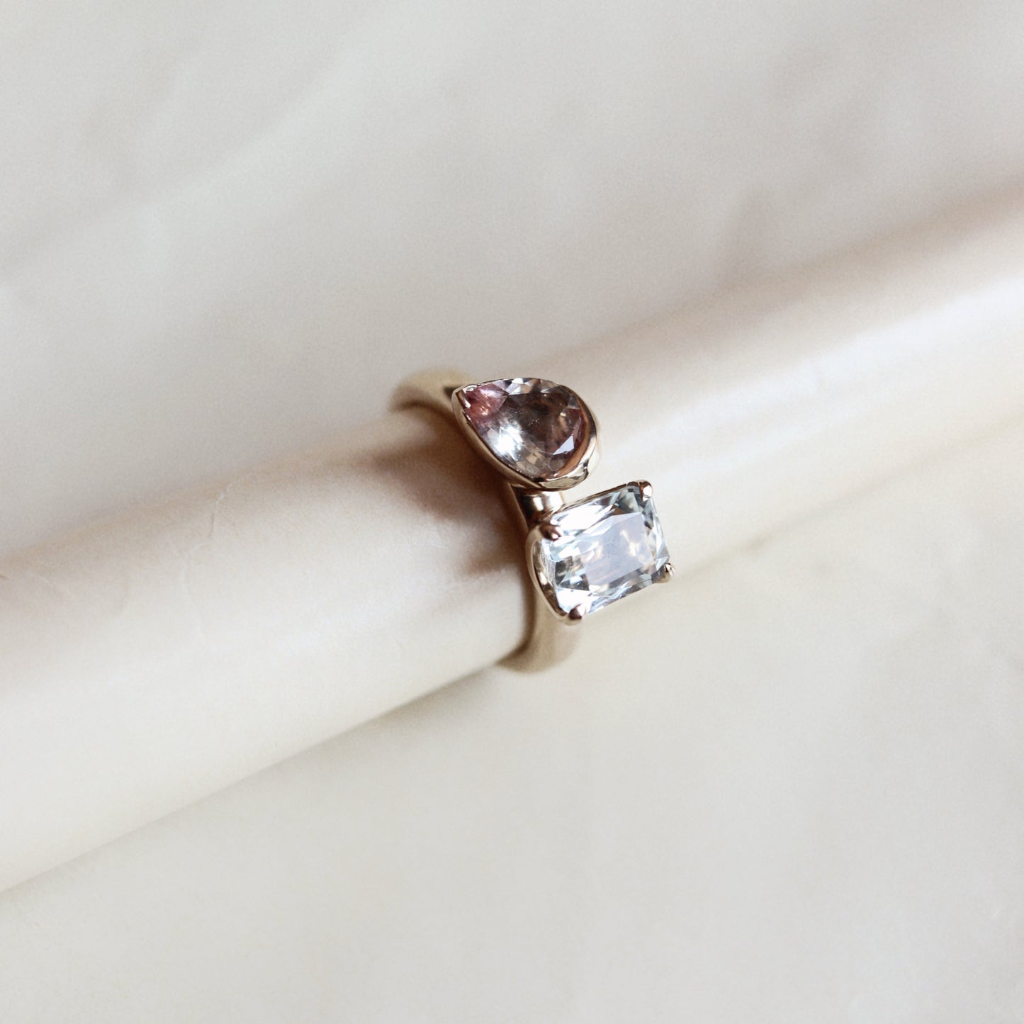 A gold ring with a marquise-cut pink gemstone and a rectangular clear gemstone, displayed on a cream-colored cylindrical stand against a light marble background.