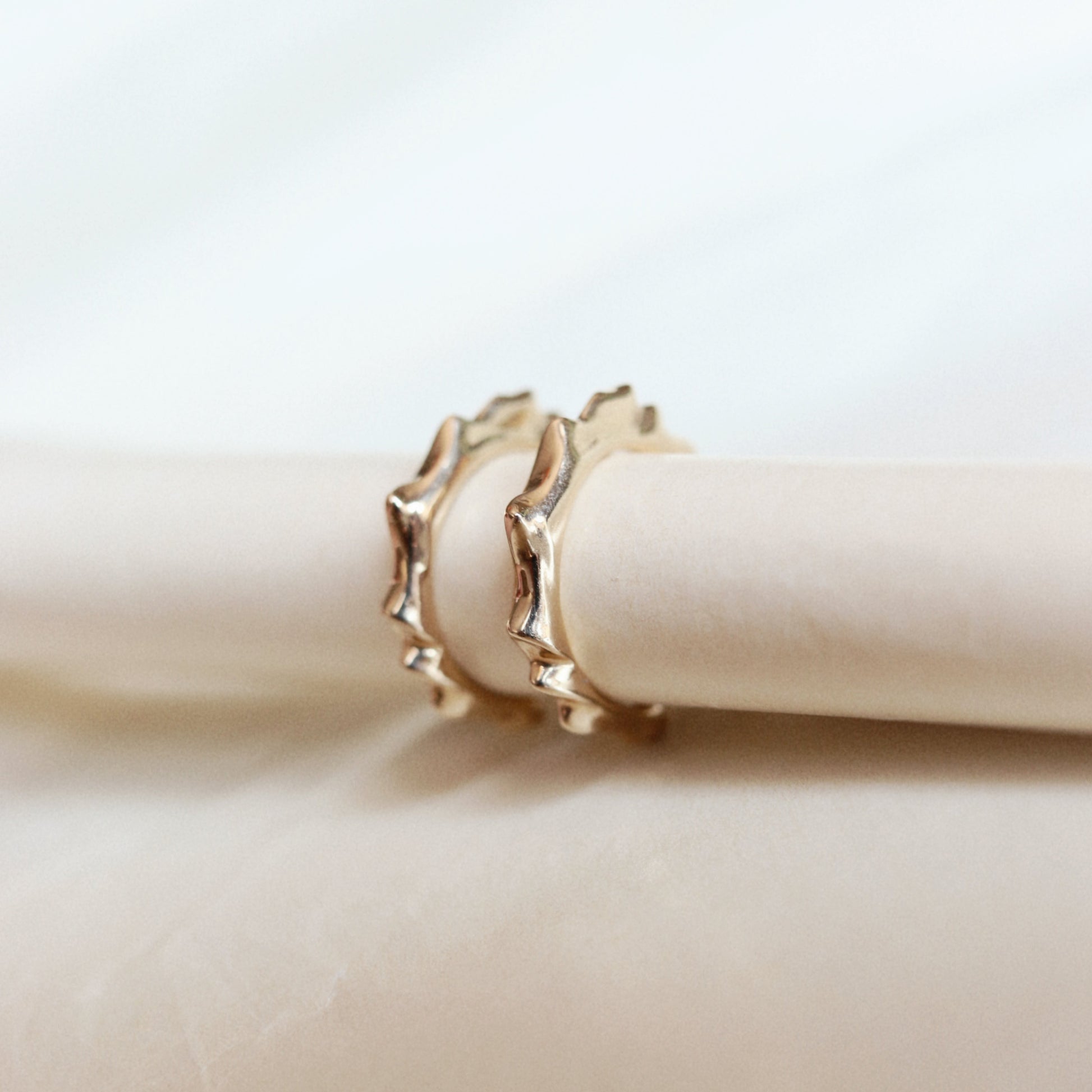 Two gold rings with a textured, wavy scalloped edge, displayed on a cream-colored cylindrical stand against a soft, light background.