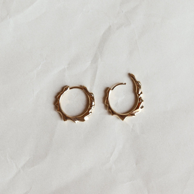 A pair of small gold hoop earrings with a textured, organic jagged edge, displayed on a wrinkled white paper background.