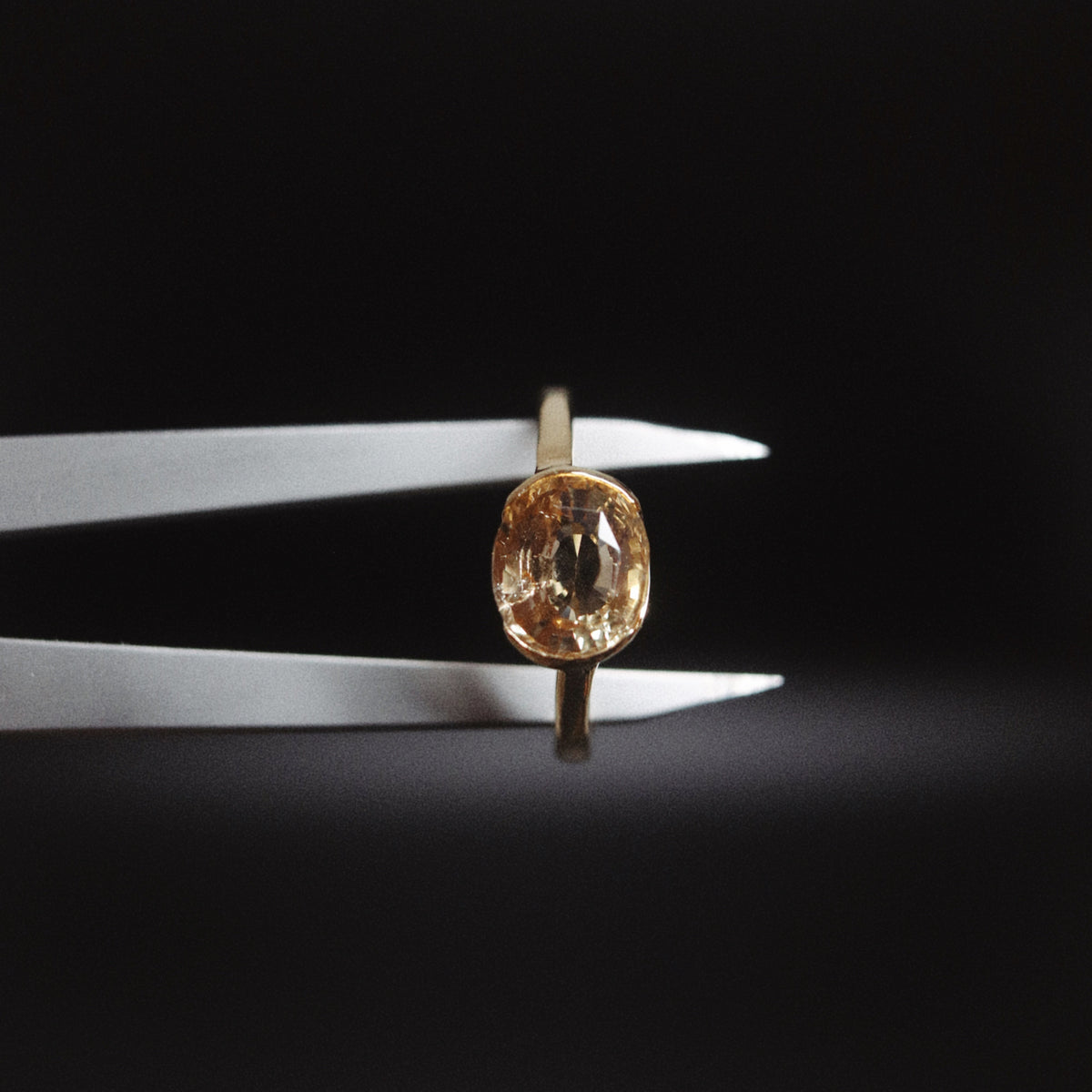 A gold ring with an oval-cut clear gemstone held in white tweezers against a dark black background.