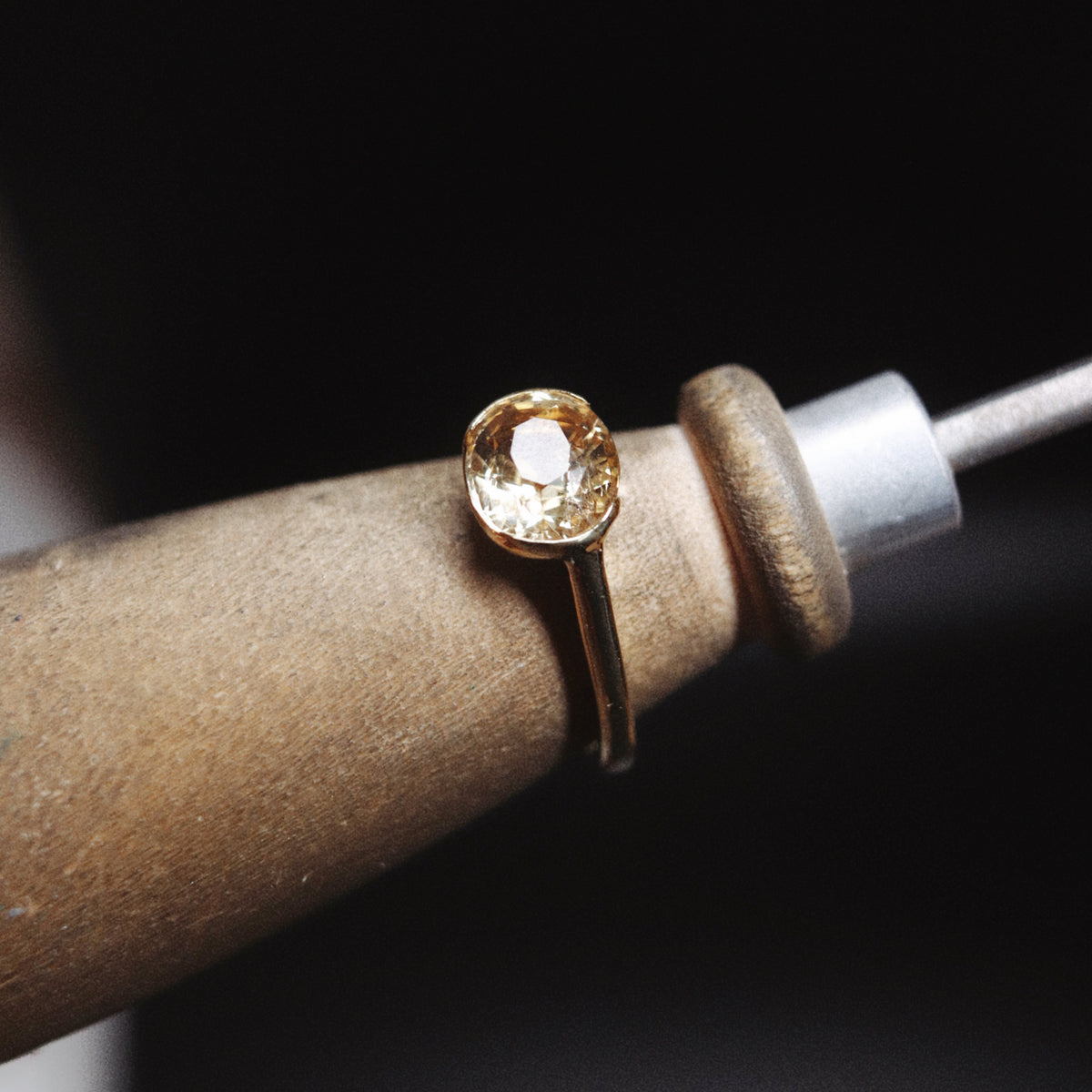 A gold ring with a cushion-cut clear gemstone held in a jeweler's loupe, set against a dark background.