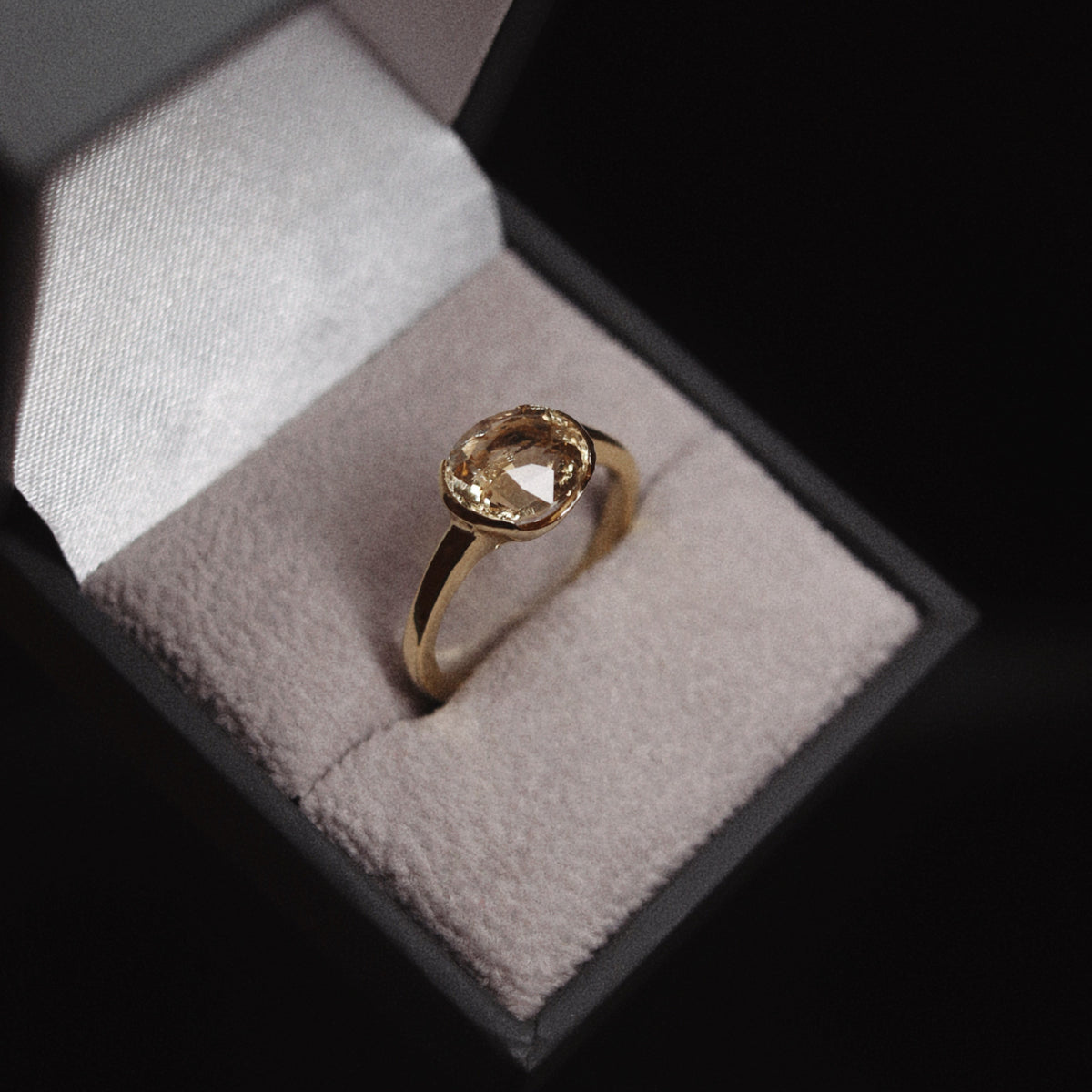 A gold ring with a round clear gemstone displayed inside an open dark gray jewelry box with a light beige velvet cushion, set against a dark background.