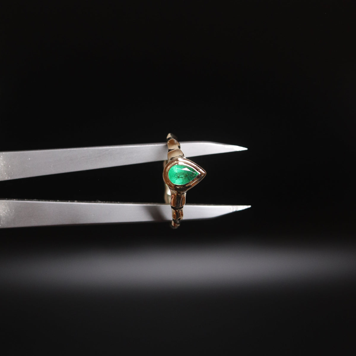 A small gold ring with a green triangular gemstone is held between metal calipers against a dark black background.