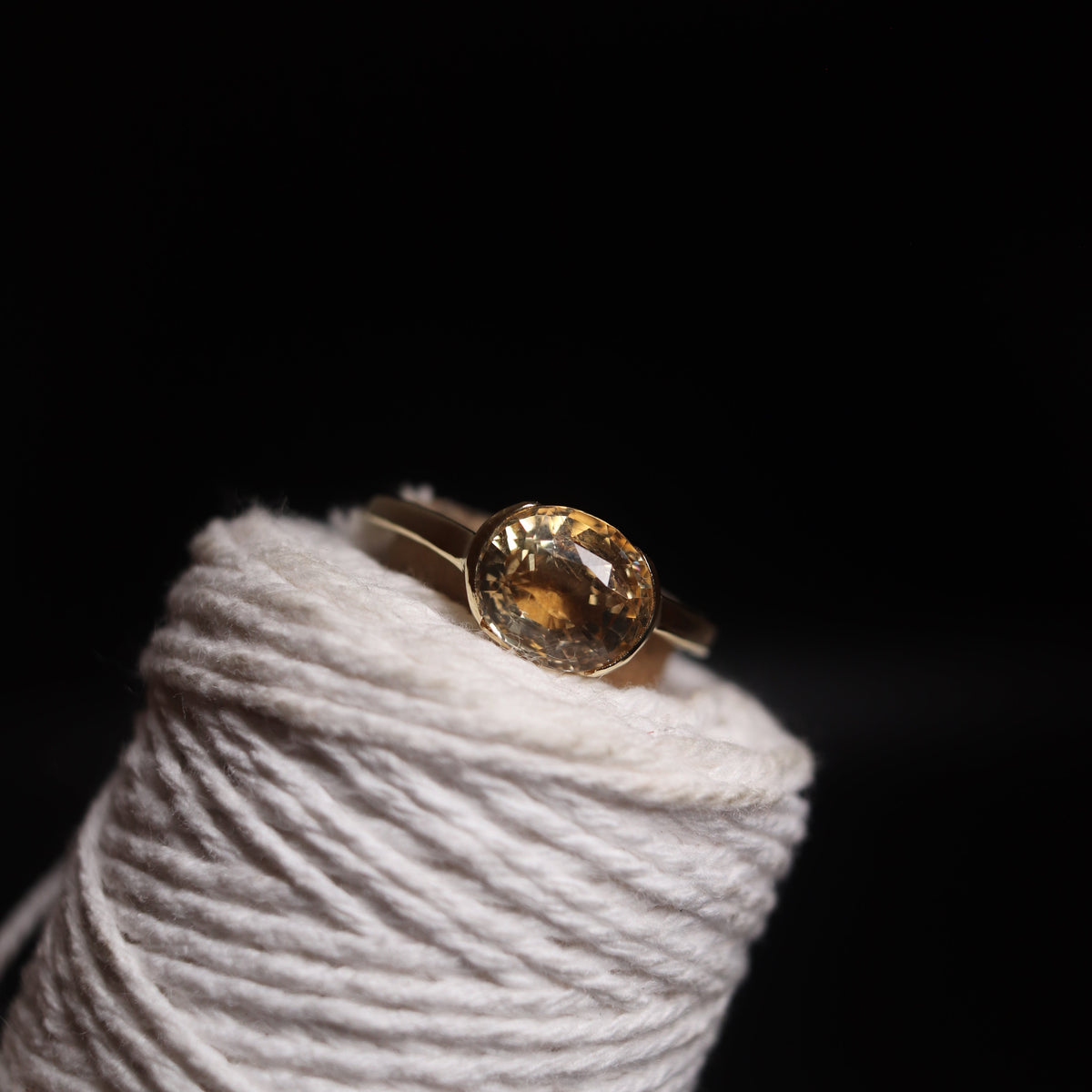 A gold ring with a round faceted amber gemstone, displayed on a spool of white thread against a solid black background.