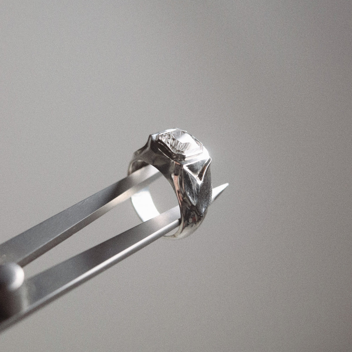A close-up of a silver ring held in metal tweezers against a soft gray background.