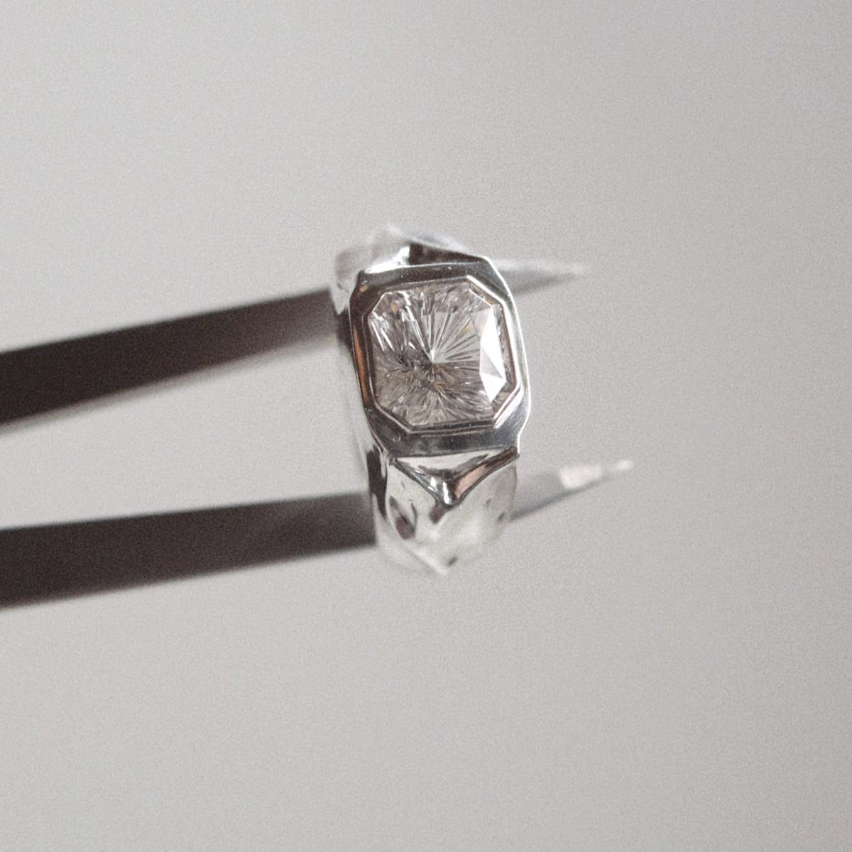 A faceted silver ring with a clear central gemstone held by black tweezers against a soft, light gray background.