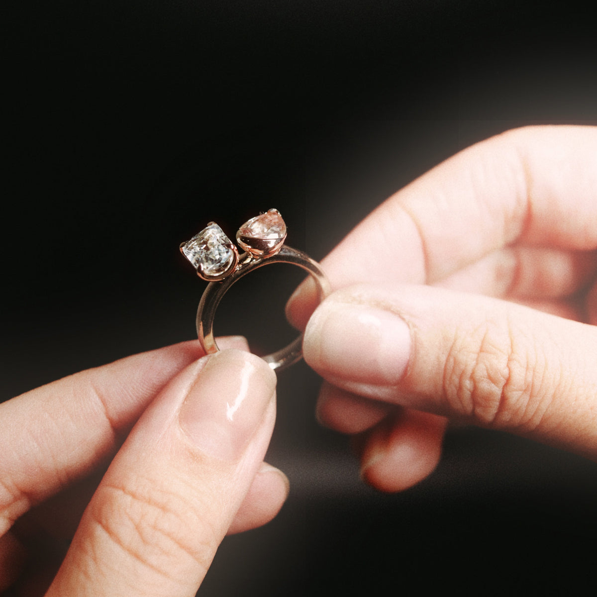 A close-up of a person's hands holding a rose gold ring with two heart-shaped gemstones against a dark, blurred background.