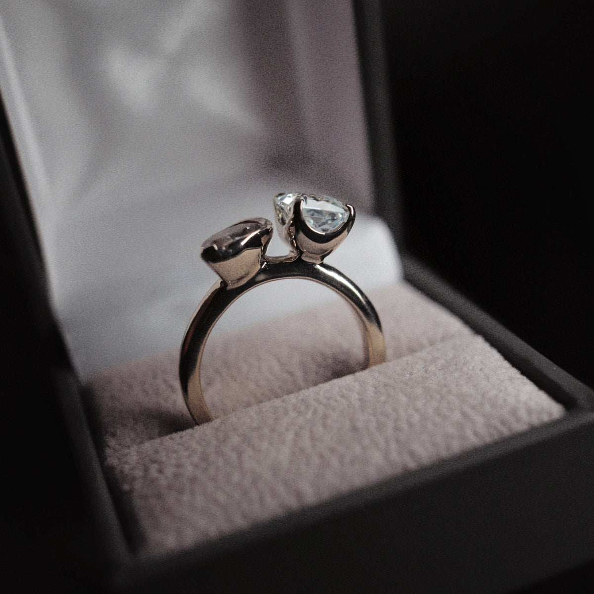 A silver ring with two heart-shaped clear gemstones displayed inside an open black jewelry box with a light pink velvet cushion.
