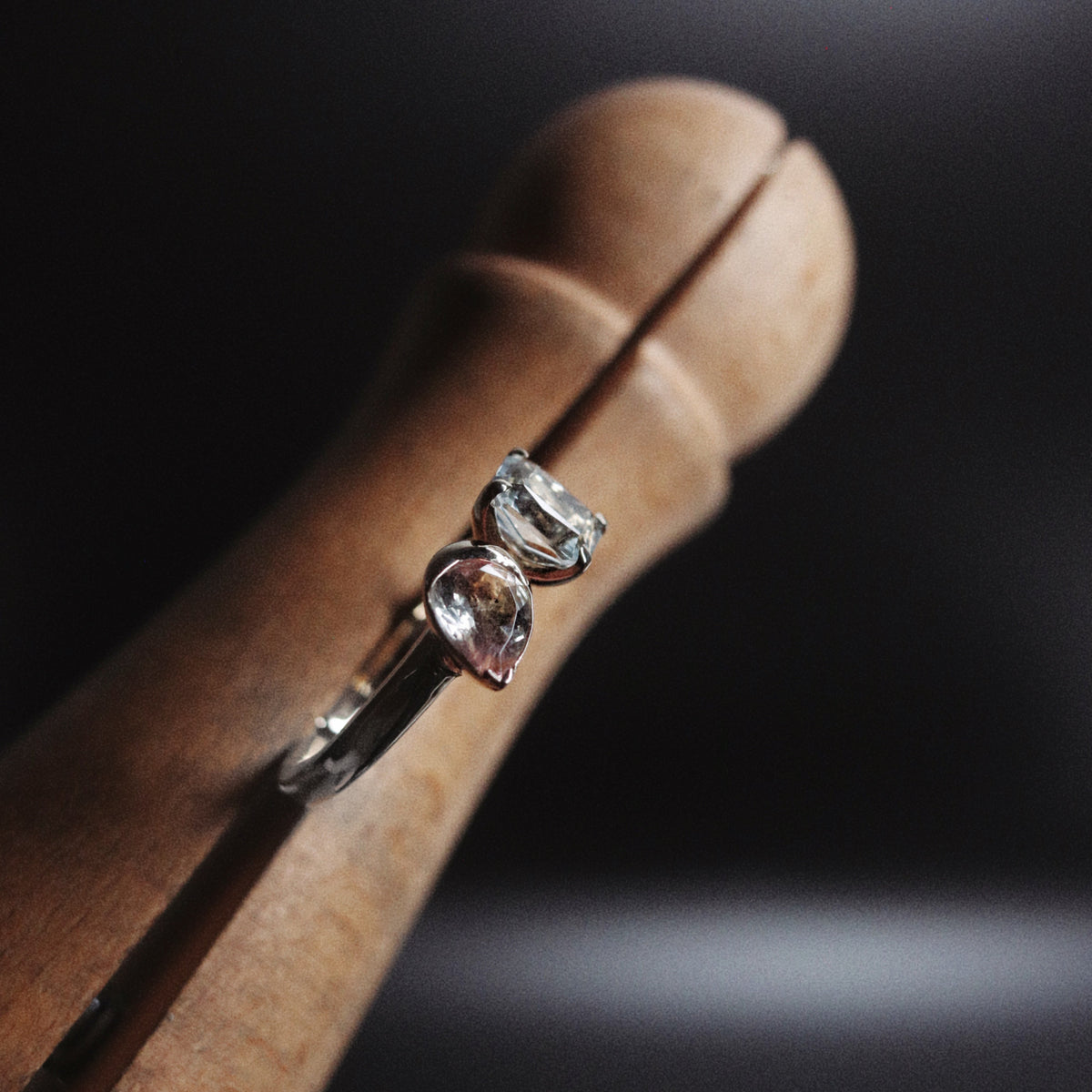 A silver ring with two clear gemstones, one marquise-cut and one round-cut, displayed on a wooden ring stand against a dark background.