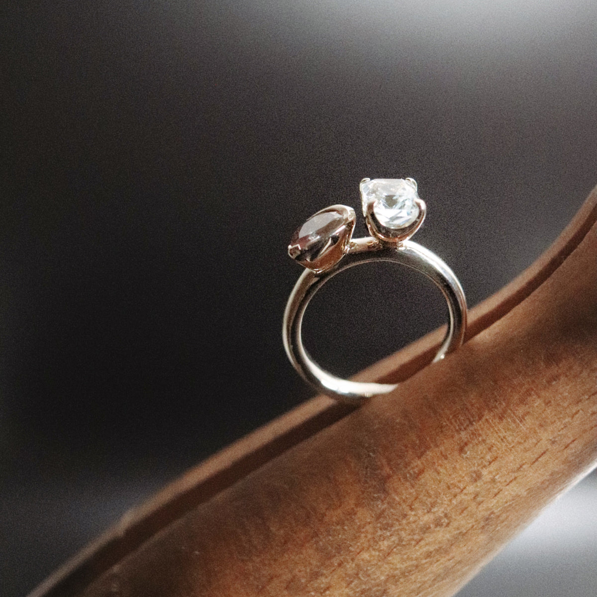 A silver ring with two clear gemstones resting on a curved wooden surface against a dark, softly lit background.