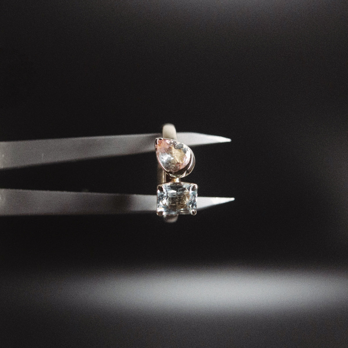A small stud earring held in metal tweezers against a dark black background. The earring features a clear rectangular gemstone below a pear-shaped pale pink gemstone in a silver-toned setting.