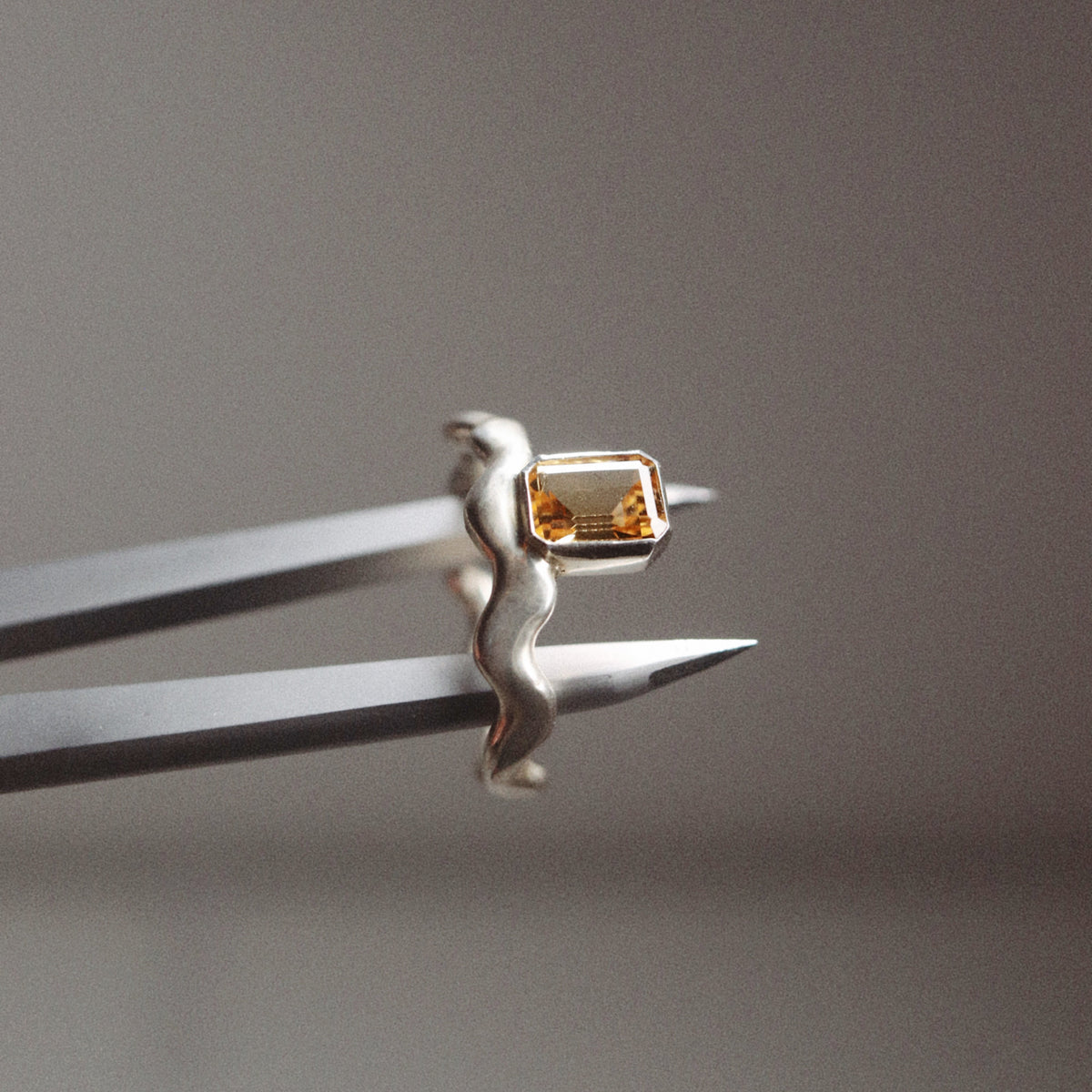 A silver ring with a wavy band and a small rectangular gold bezel containing an amber gemstone, held in silver tweezers against a neutral gray background.