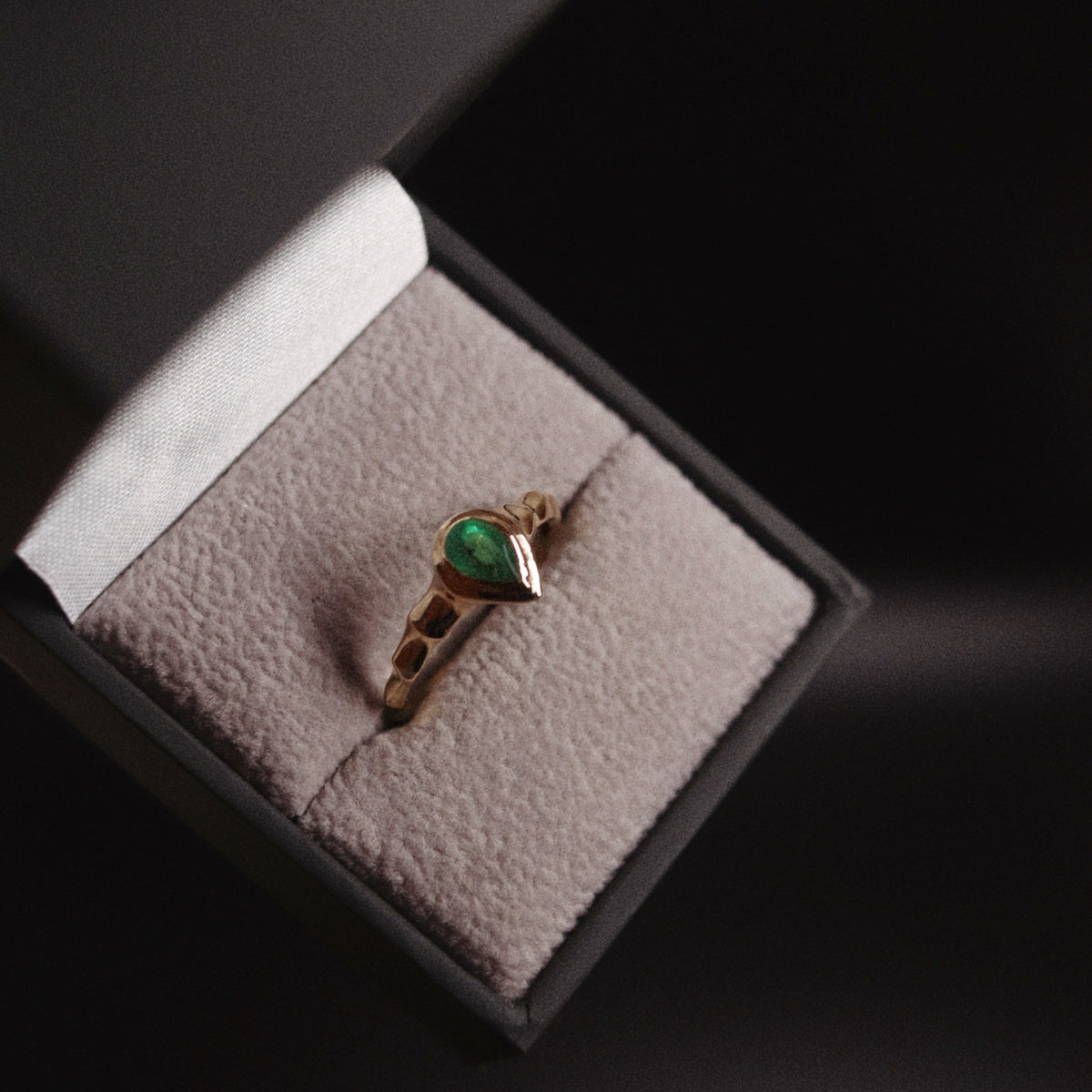 A gold ring with a green heart-shaped gemstone displayed on a textured gray ring box, set against a dark background.