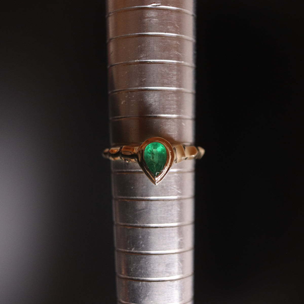 A gold ring with a teardrop-shaped green emerald gemstone, displayed on a silver ring stand against a dark background.