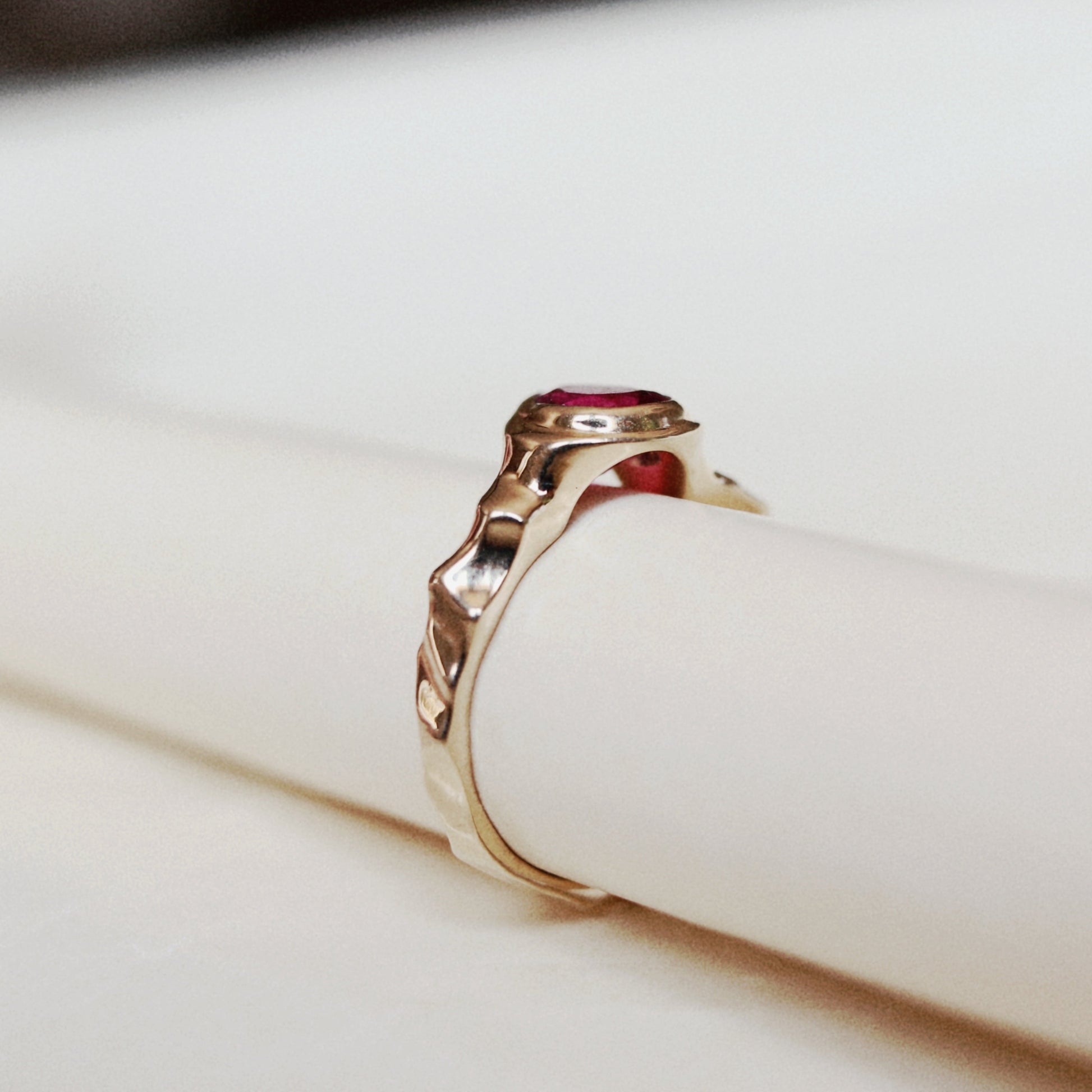 A gold ring with a textured, organic-shaped band and a small round red gemstone, displayed on a white cylindrical stand.