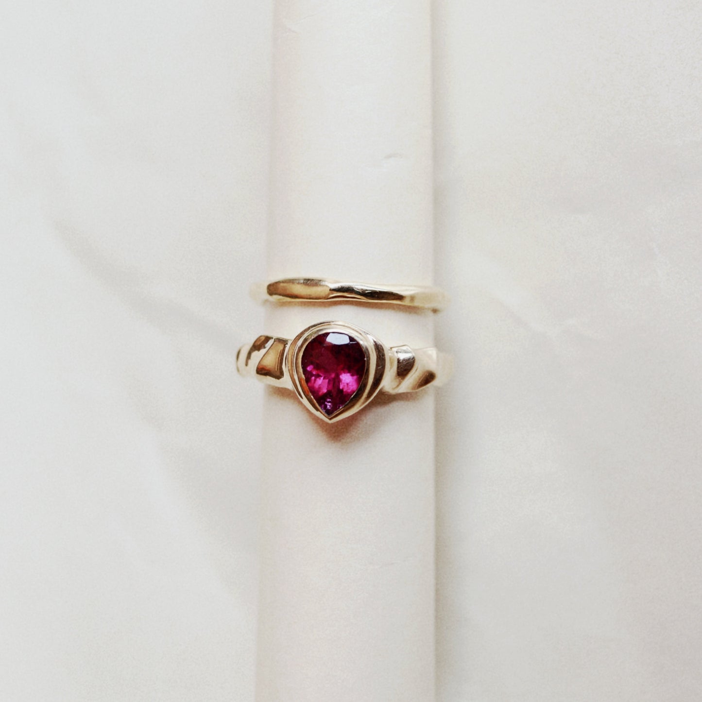 A gold ring with a heart-shaped pink gemstone, displayed on a cream-colored ring holder against a light marble background.