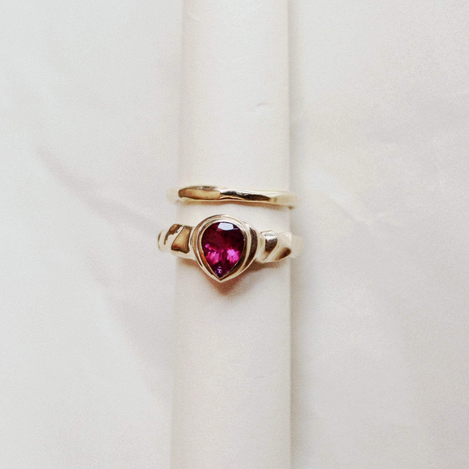 A gold ring with a heart-shaped pink gemstone, displayed on a cream-colored ring holder against a light marble background.
