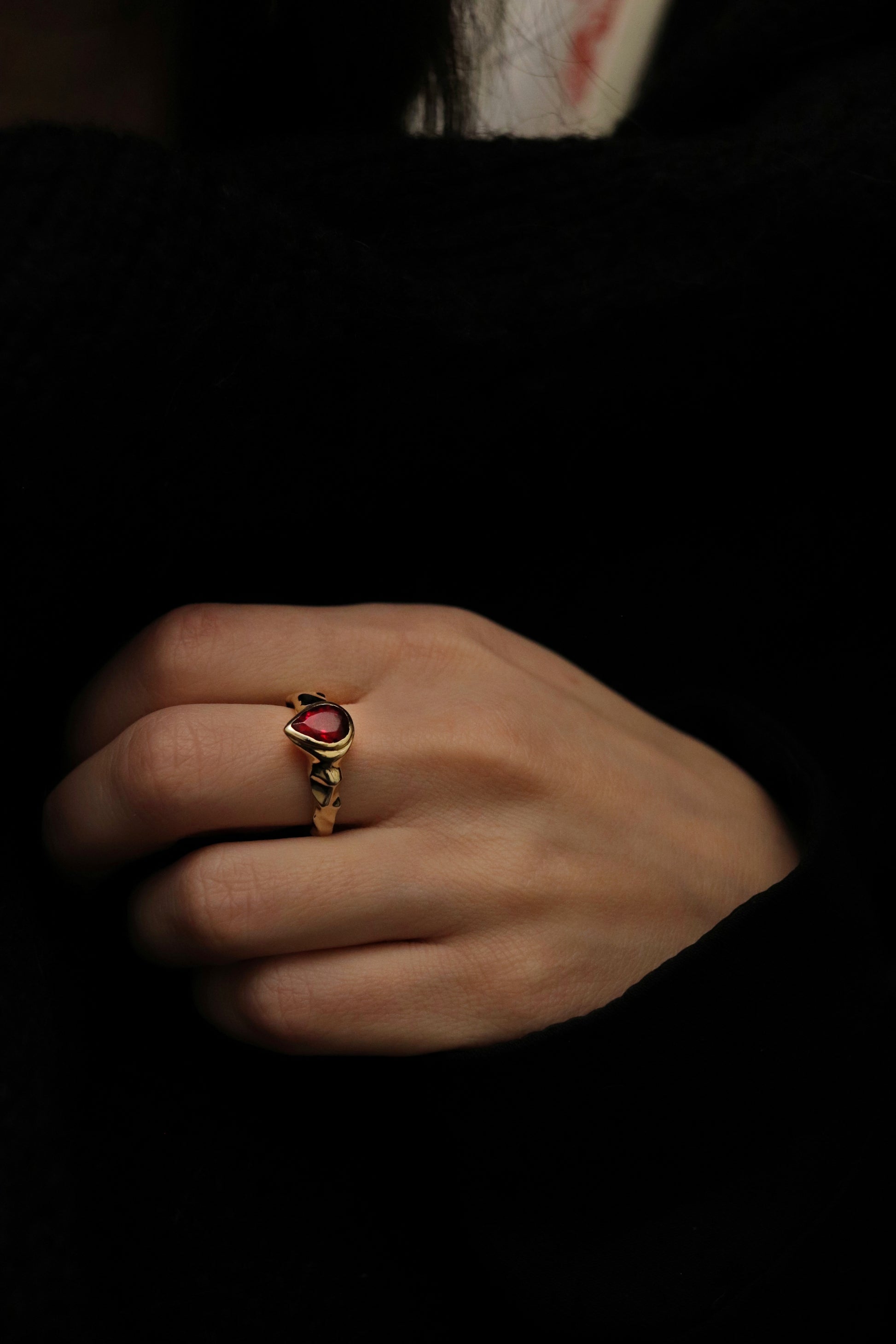 Hand wearing a gold ring with a red stone on a dark background