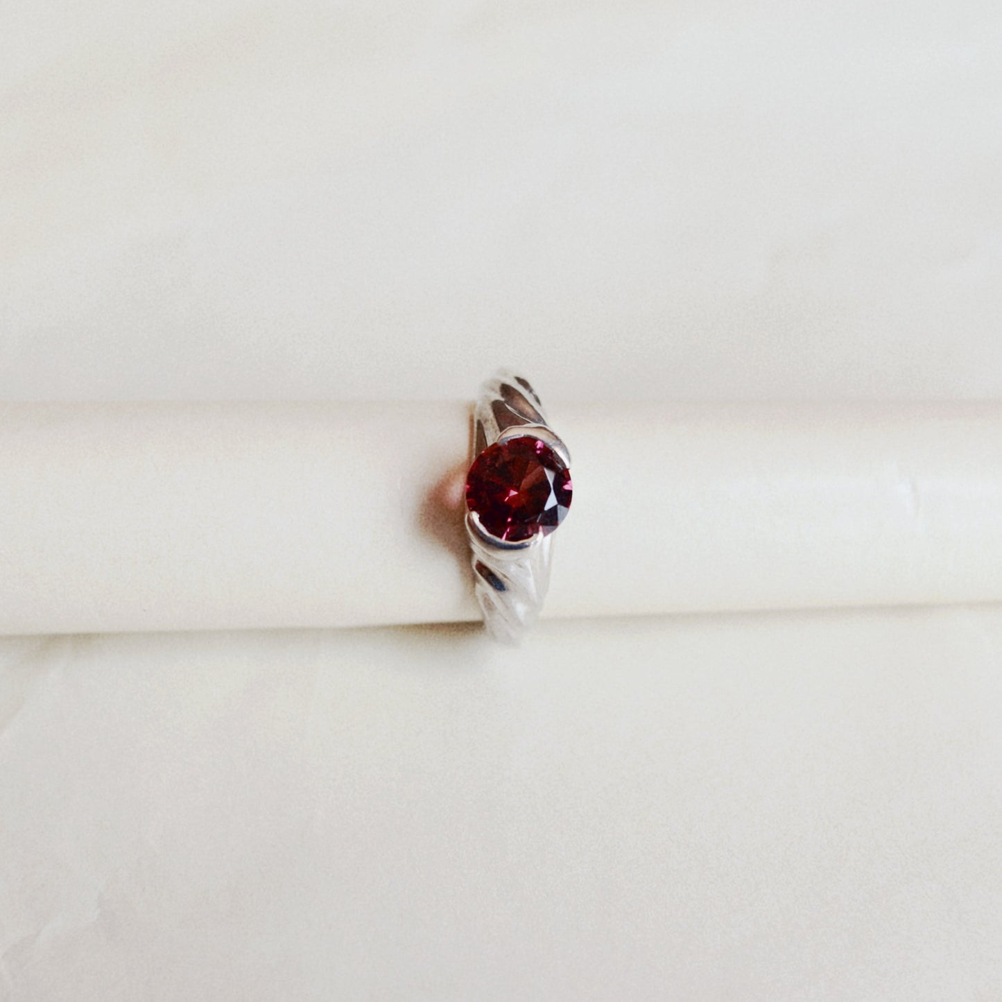 A silver ring with a round-shaped dark red gemstone, displayed on a white cylindrical stand against a soft, light beige background.