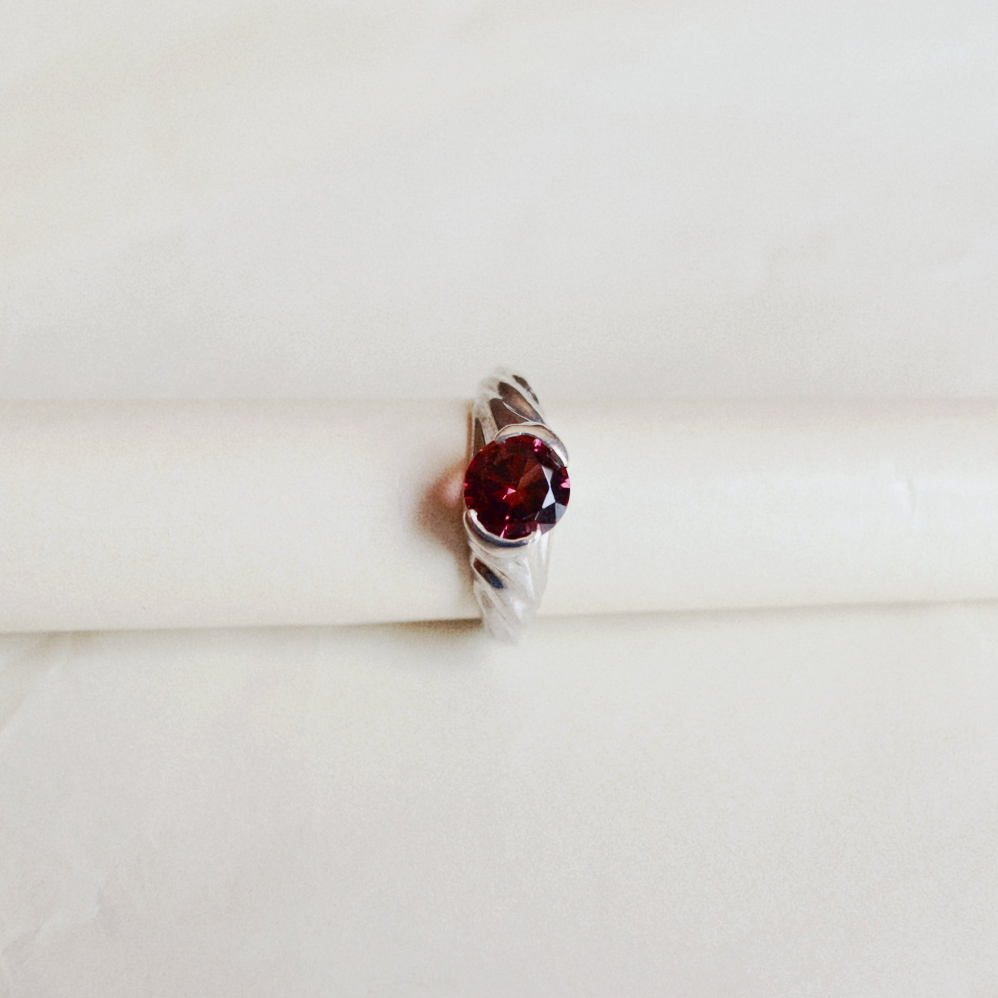 A silver ring with a round-shaped dark red gemstone, displayed on a white cylindrical stand against a soft, light beige background.