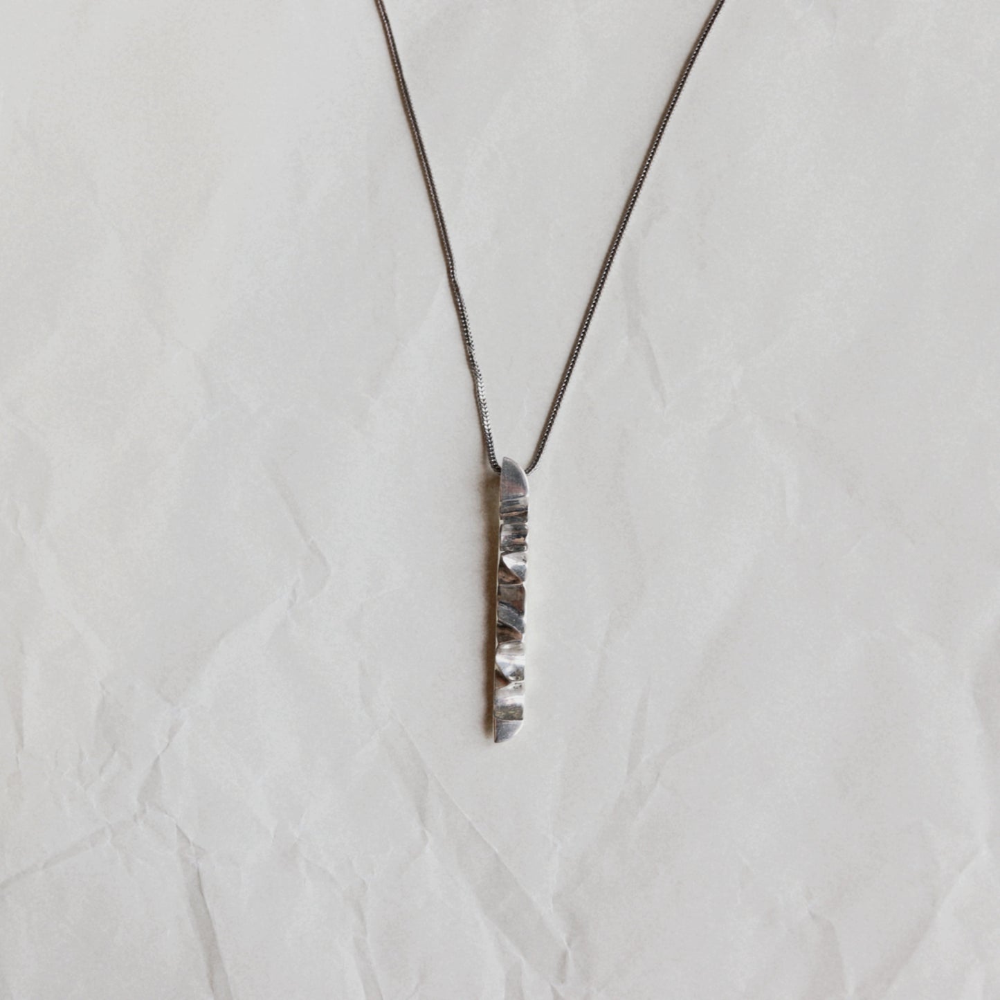 A silver-toned vertical bar pendant necklace with a textured, hammered finish on a matching fine chain, displayed on a crumpled off-white paper background.