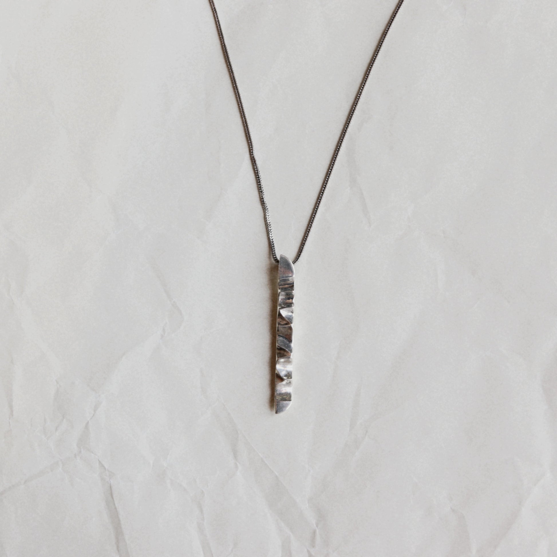 A silver-toned vertical bar pendant necklace with a textured, hammered finish on a matching fine chain, displayed on a crumpled off-white paper background.