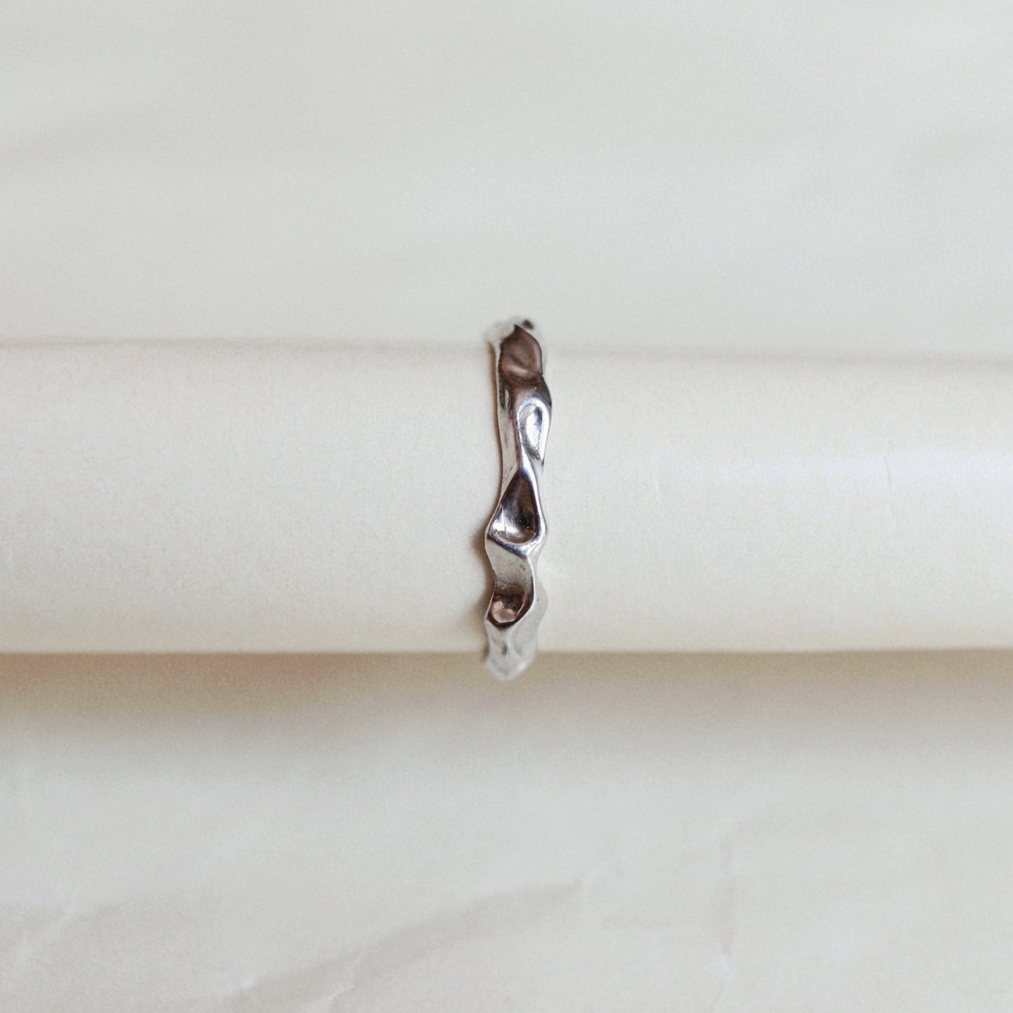 A slim, wavy sterling silver ring displayed on a cream-colored ring holder against a light beige background.