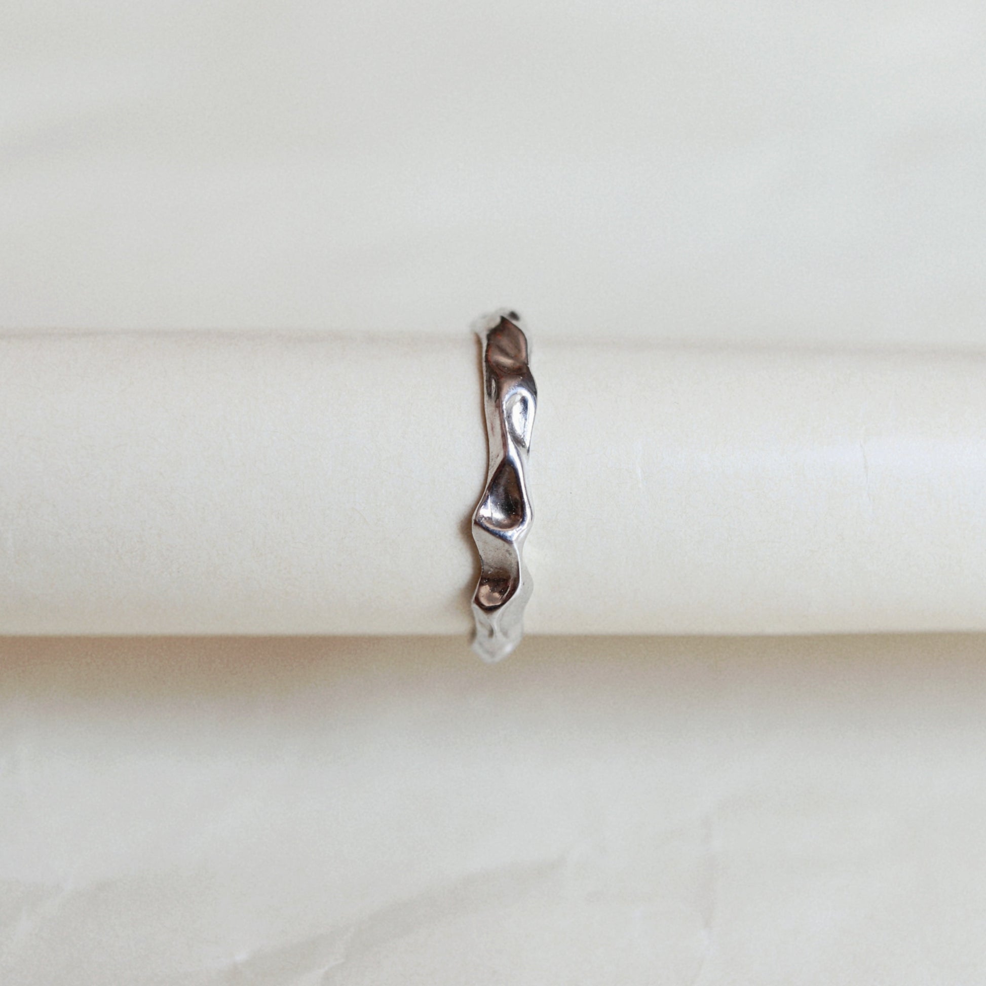 A slim, wavy sterling silver ring displayed on a cream-colored ring holder against a light beige background.