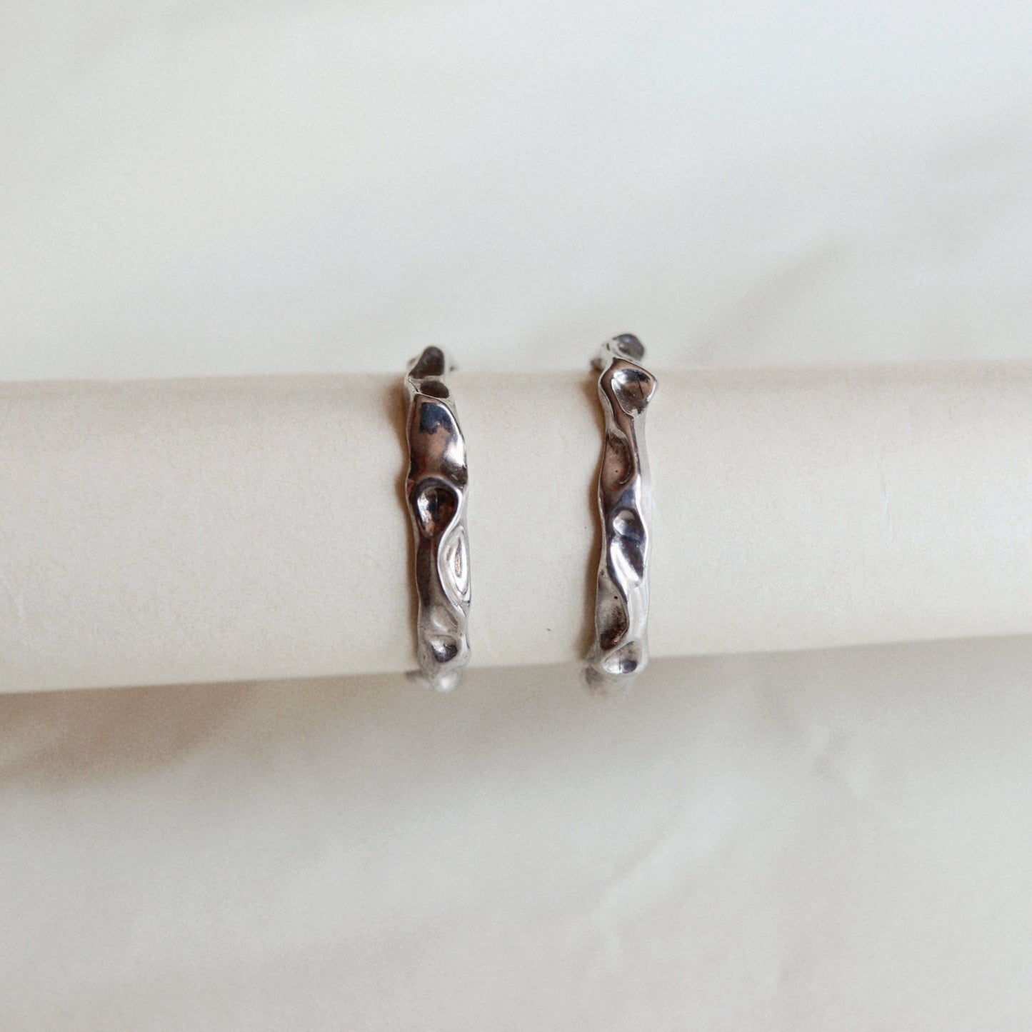 A pair of silver huggie hoop earrings with a wavy, organic textured design, displayed on a white paper ring holder against a light beige marble background.