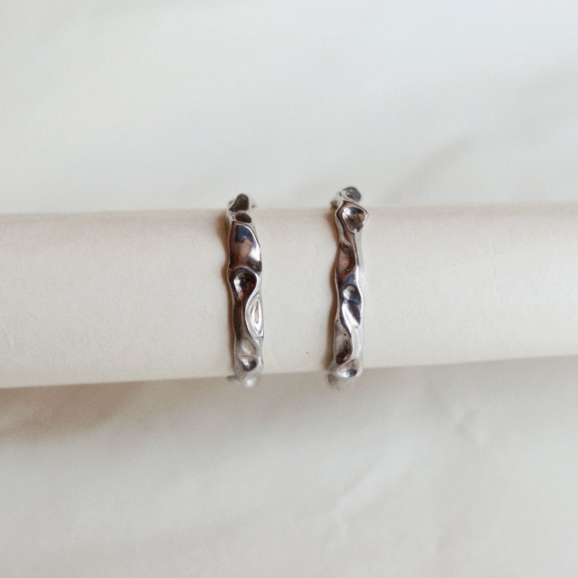 A pair of silver huggie hoop earrings with a wavy, organic textured design, displayed on a white paper ring holder against a light beige marble background.