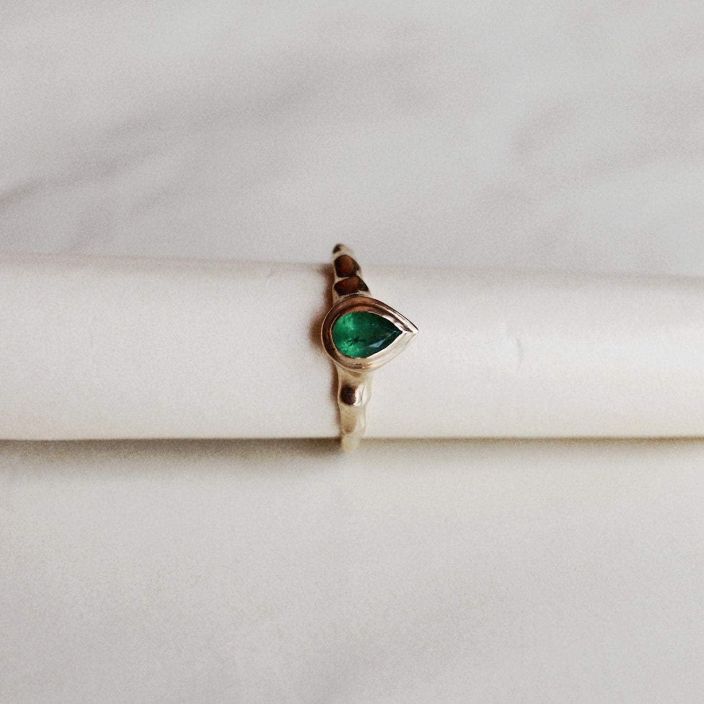 A gold ring featuring a teardrop-shaped green gemstone, displayed on a white cylindrical stand against a light marble background.