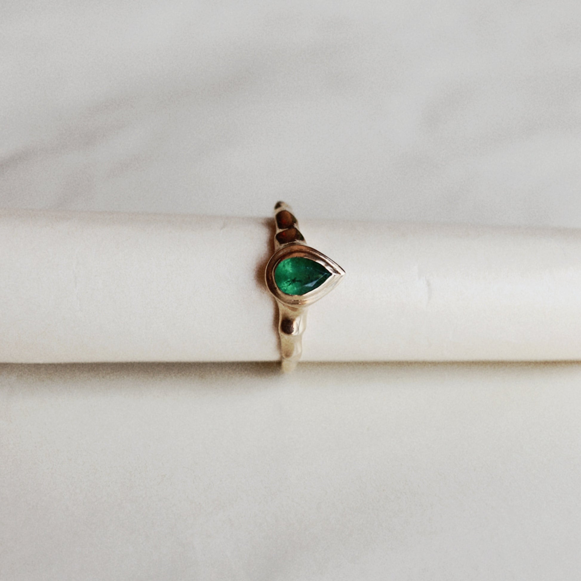 A gold ring featuring a teardrop-shaped green gemstone, displayed on a white cylindrical stand against a light marble background.