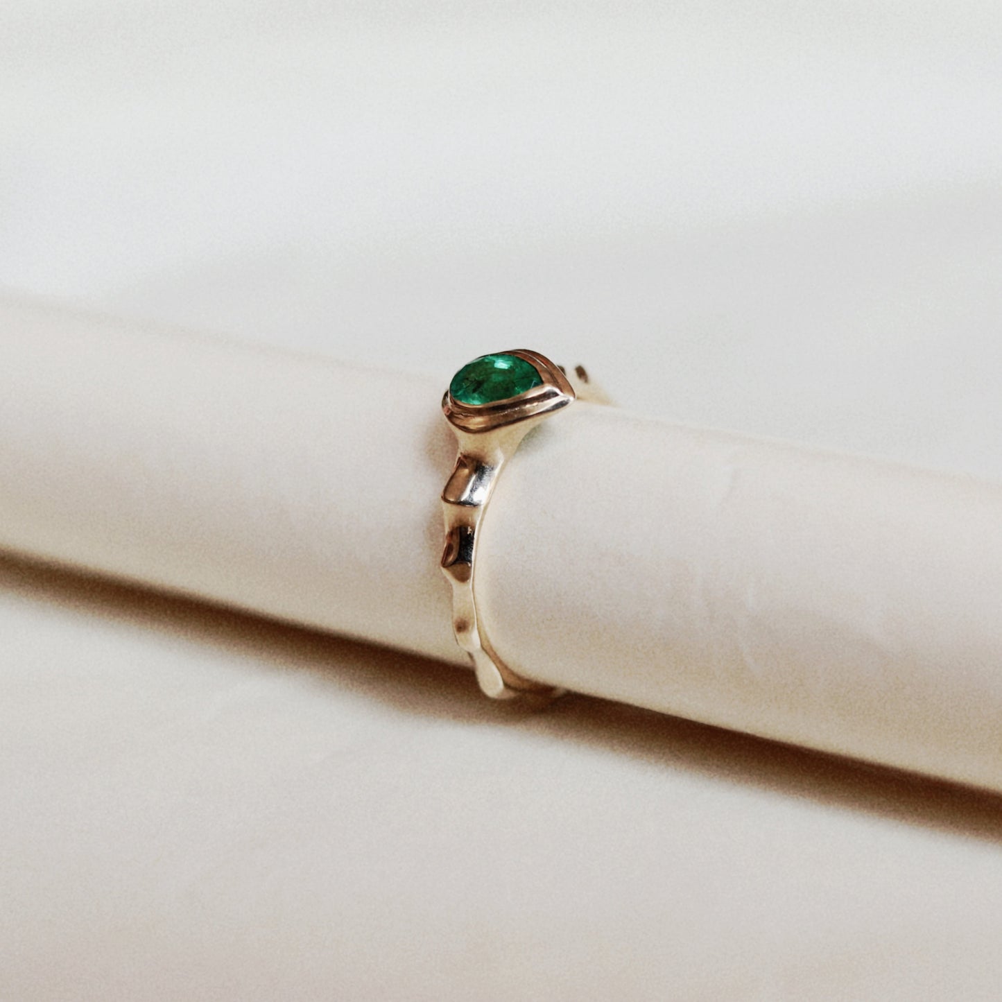A gold ring with a green oval gemstone, displayed on a white cylindrical ring stand against a light beige background.