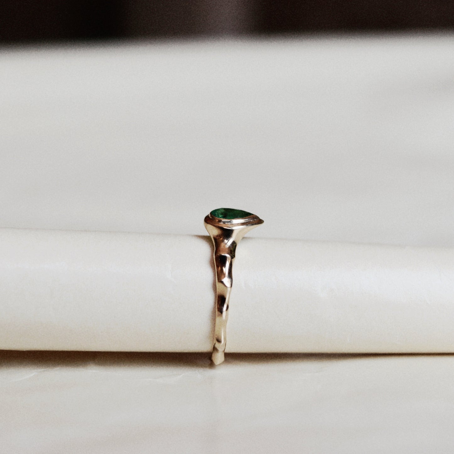 A gold ring with a small green teardrop gemstone, displayed on a white cylindrical stand.
