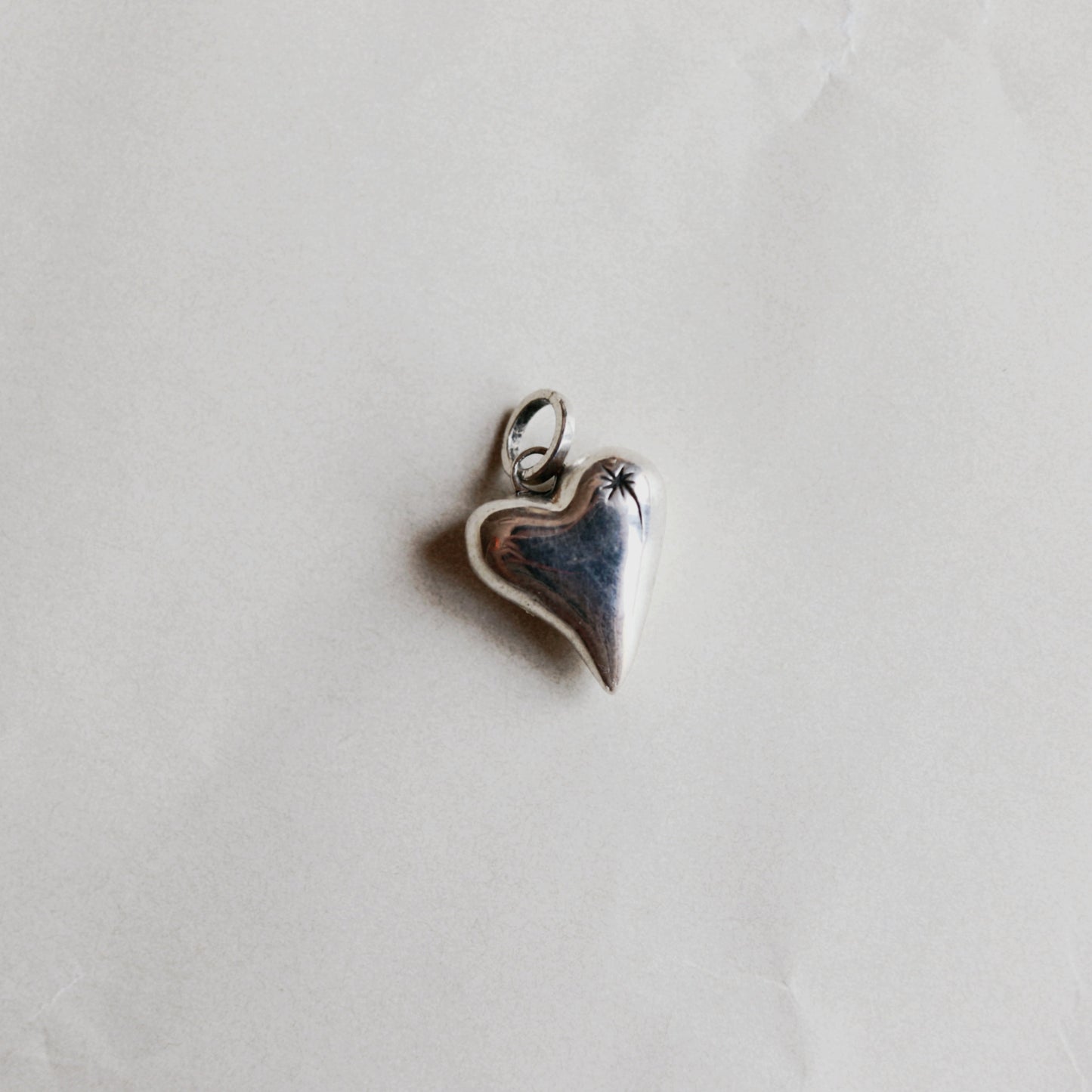 A small heart-shaped pendant charm with a silver-toned border and bail, featuring a dark blue center with a tiny white star detail, resting on a light gray textured surface.