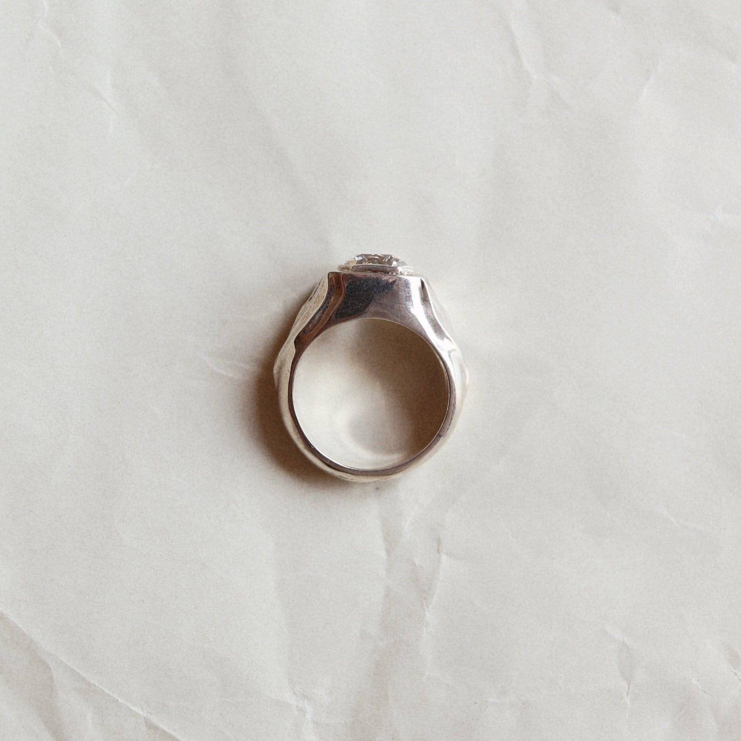 A silver-toned ring with a textured, hammered band and a small clear stone set in the center, resting on a crumpled white paper background.