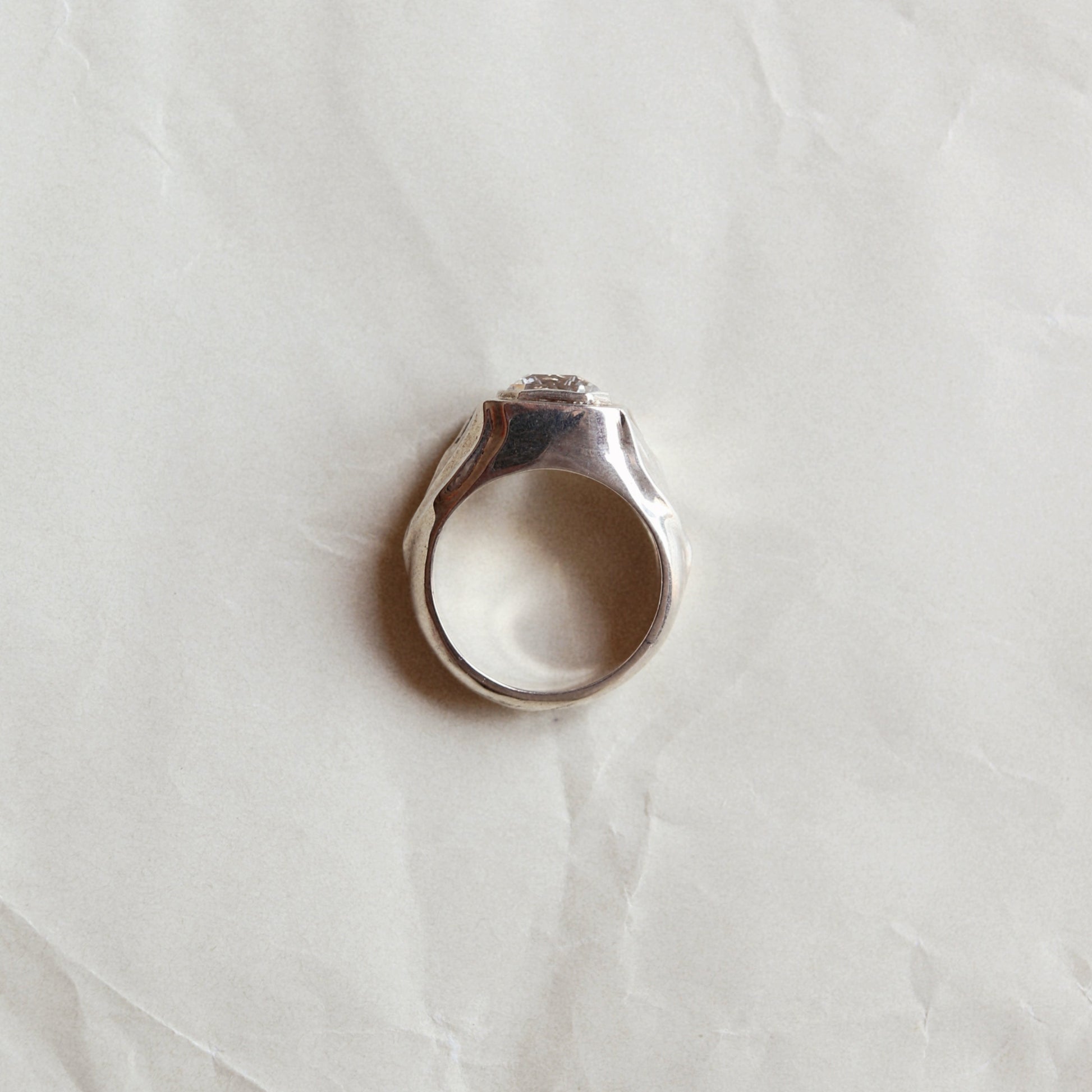 A silver-toned ring with a textured, hammered band and a small clear stone set in the center, resting on a crumpled white paper background.