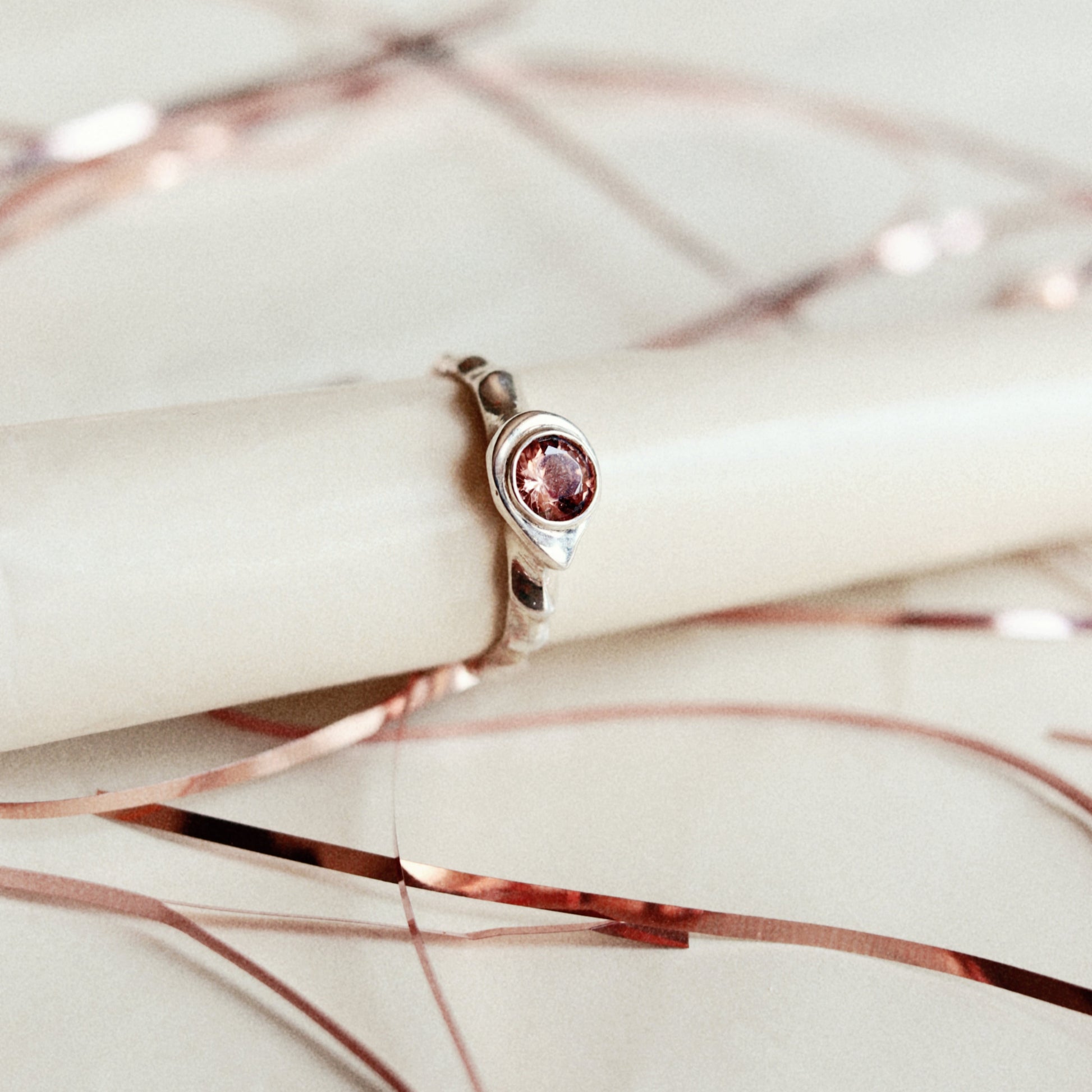A rose gold ring with a marquise-cut pink gemstone, displayed on a cream-colored cylindrical stand with rose gold wire accents on a light beige background.