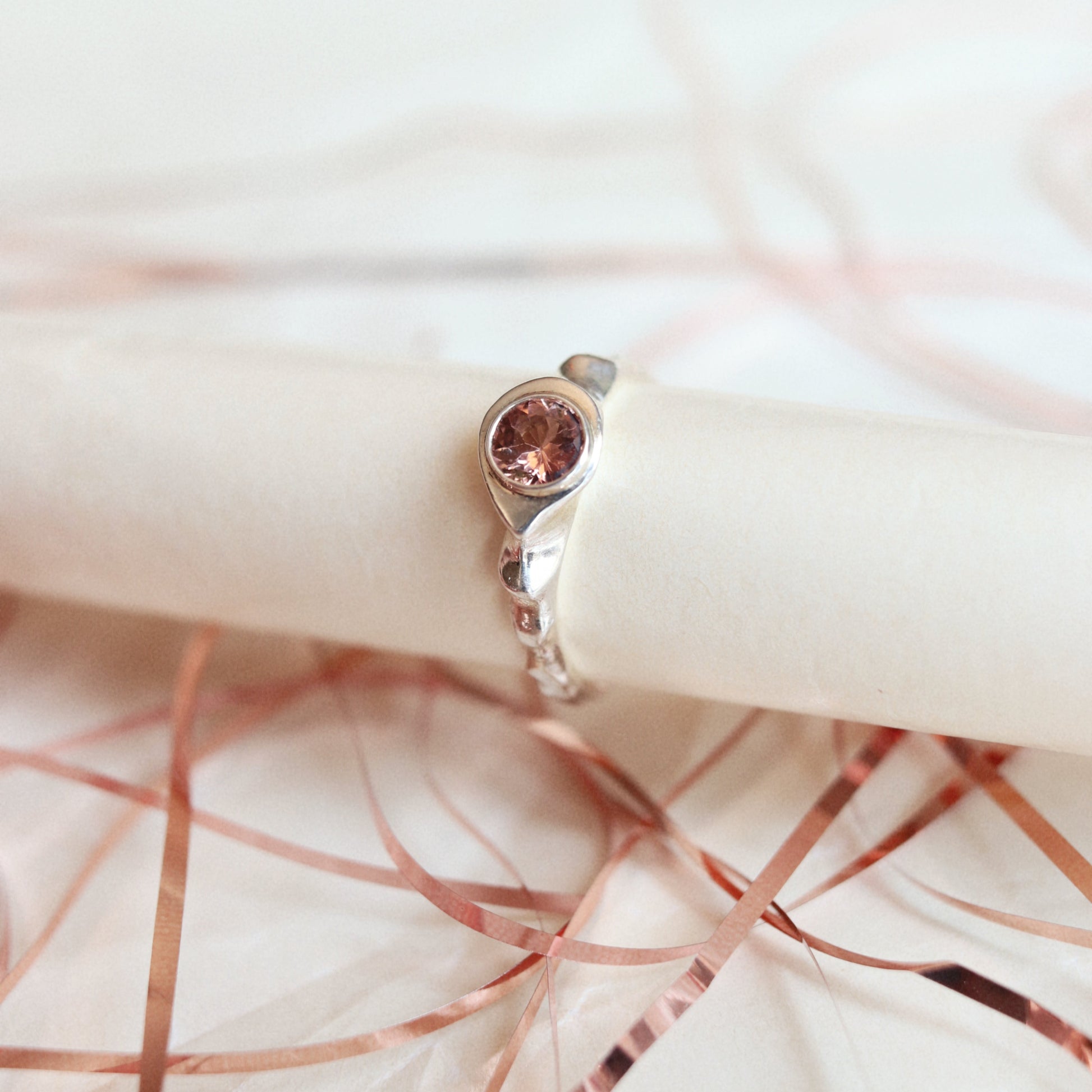 A silver ring with a round rose gold gemstone, displayed on a cream-colored cylindrical stand with decorative rose gold ribbon strands in the background.