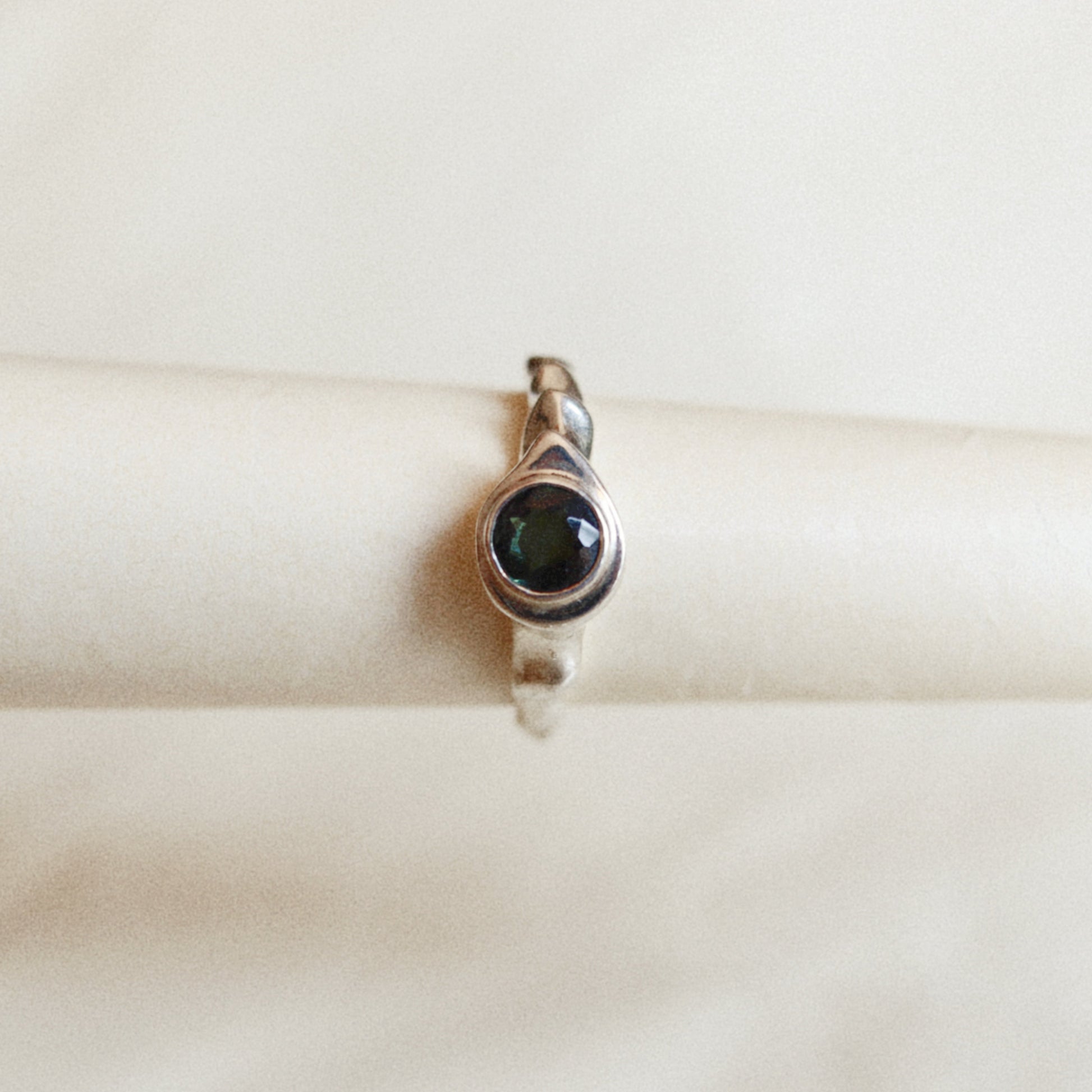 A sterling silver ring with a dark green iridescent cabochon gemstone, displayed on a cream-colored ring stand against a soft, light beige background.