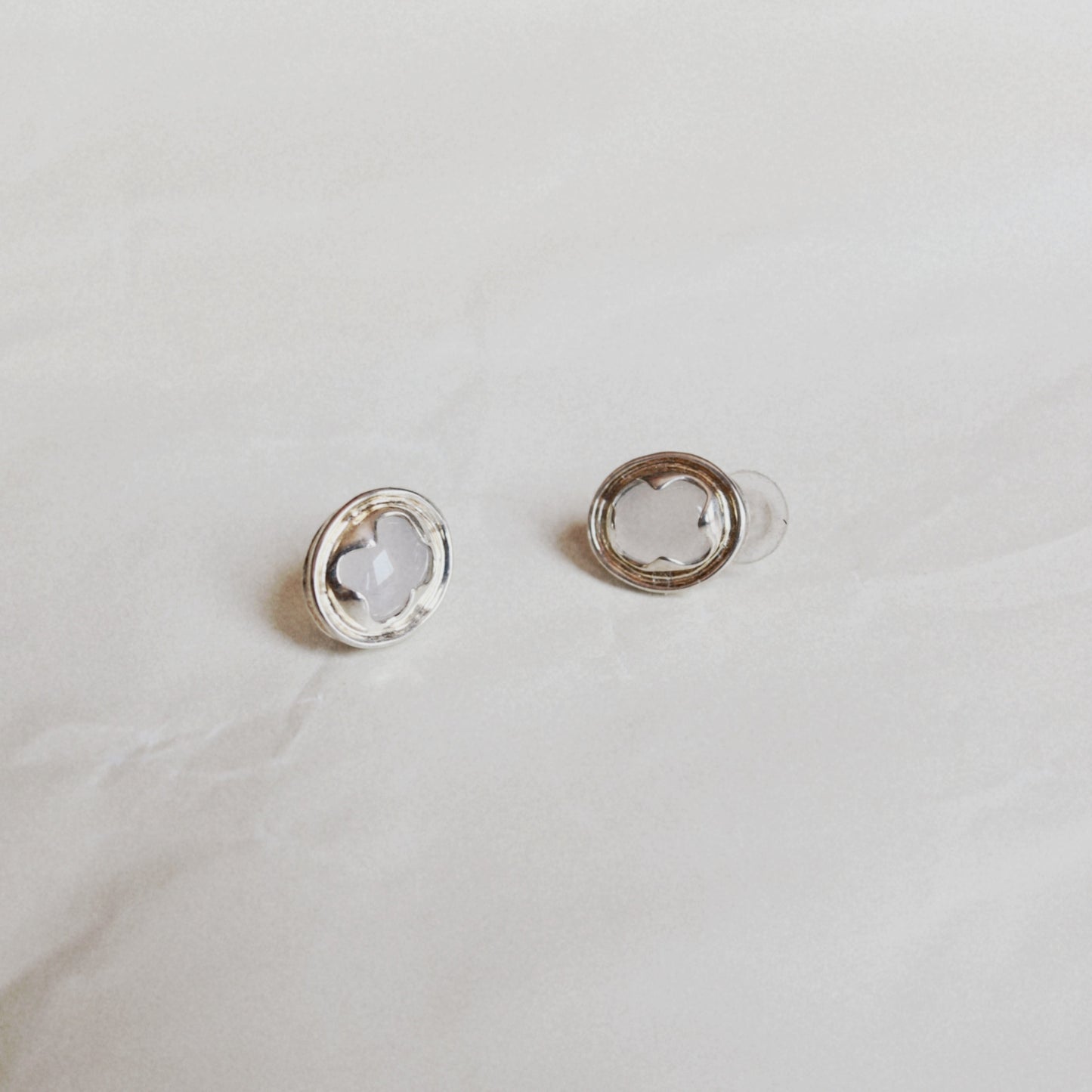 A pair of silver-toned stud earrings with a circular outer ring and a smaller inner ring holding a clear, flower-shaped stone, displayed on a light textured surface.