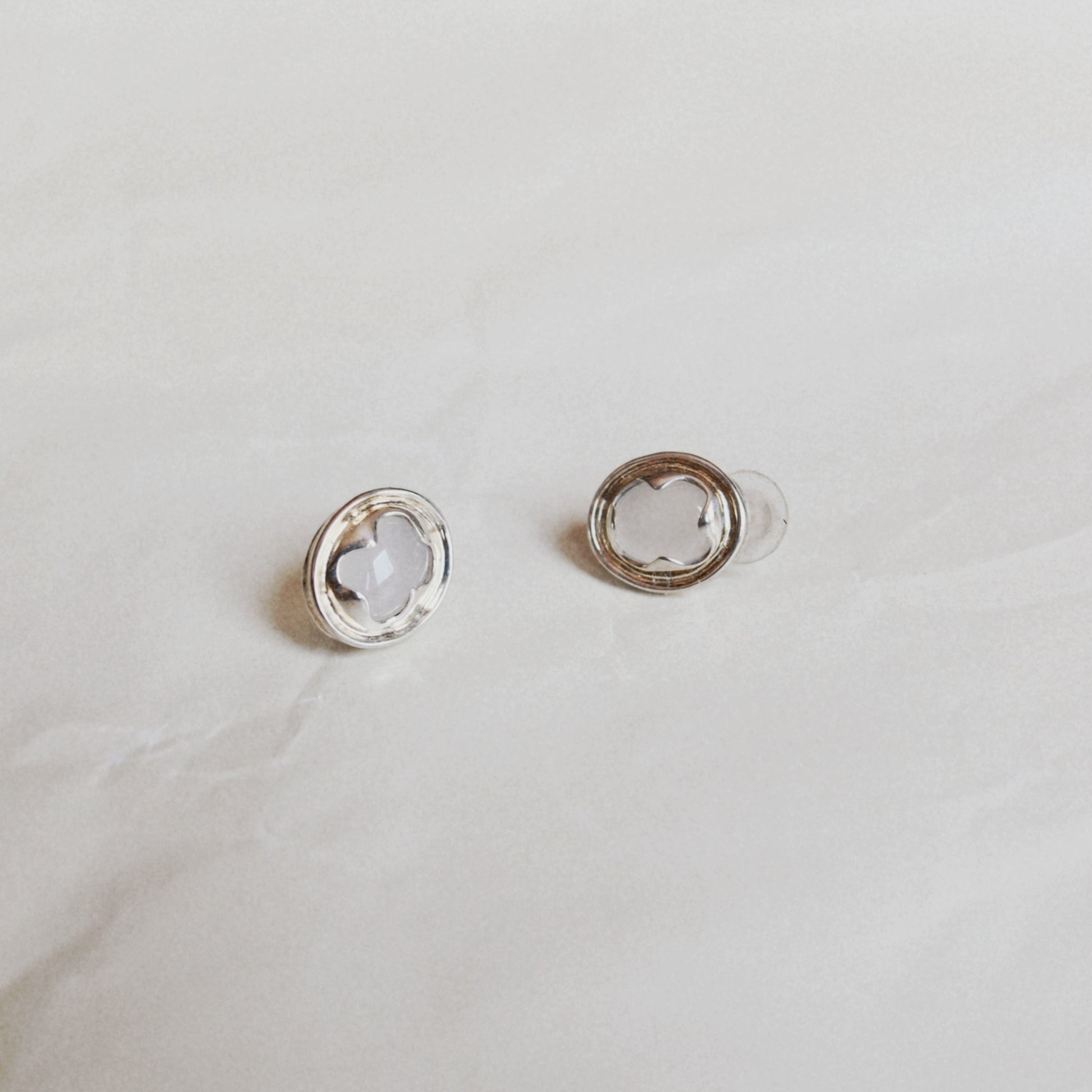 A pair of silver-toned stud earrings with a circular outer ring and a smaller inner ring holding a clear, flower-shaped stone, displayed on a light textured surface.