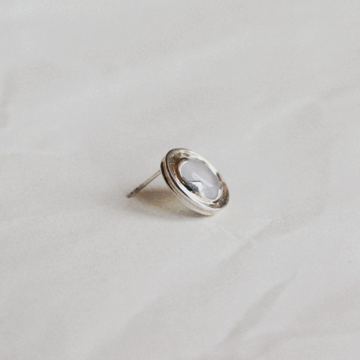 A silver-toned oval stud earring with a milky white cabochon gemstone, photographed on a soft, light gray textured fabric background.