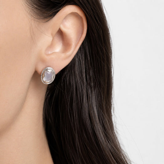 Close-up of an ear wearing a silver earring with a clear gemstone on a neutral background