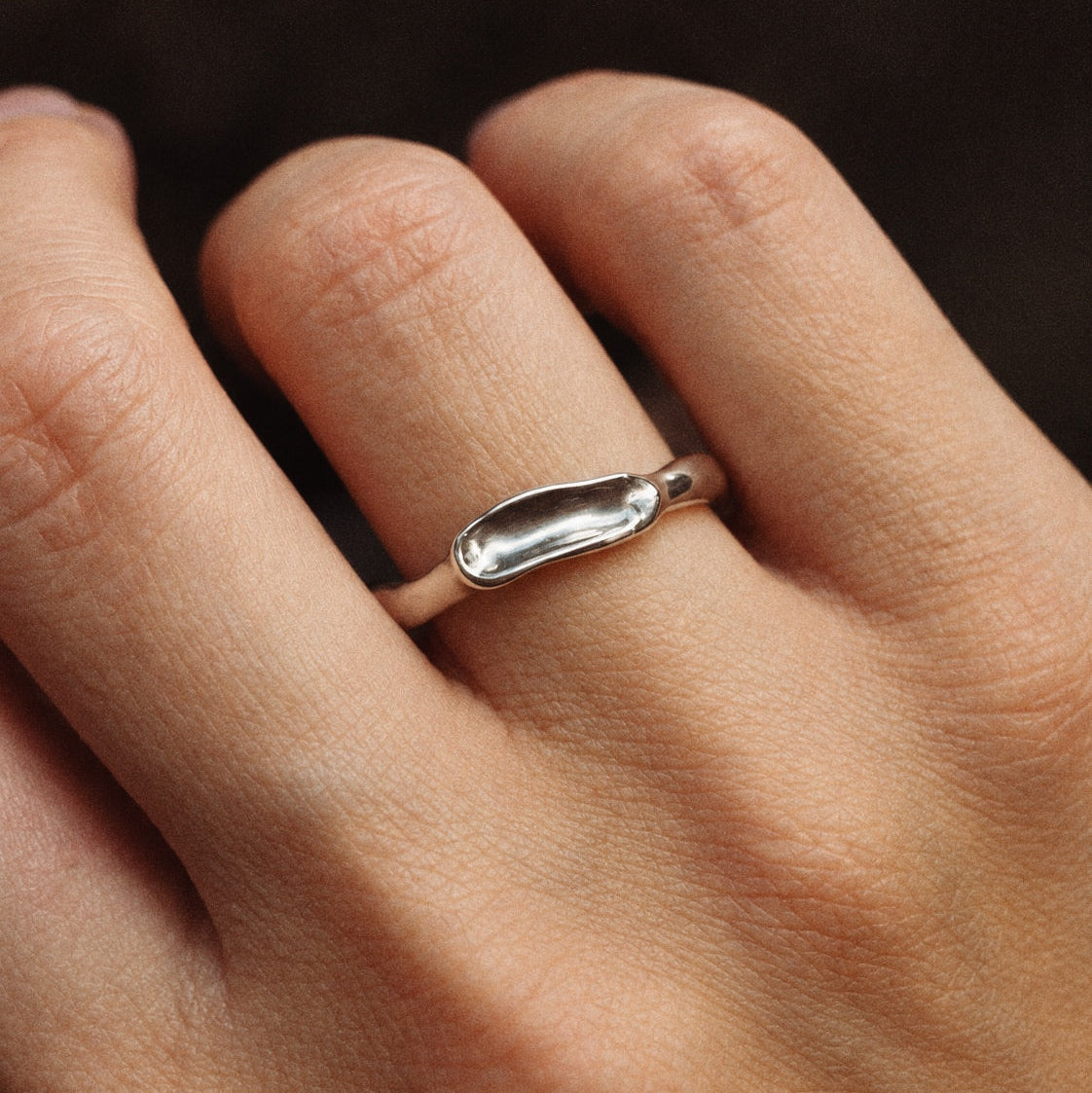 A close-up of a hand wearing a minimalist silver ring with a smooth, elongated oval design on the ring finger.