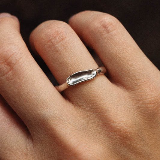 A close-up of a hand wearing a minimalist silver ring with a smooth, elongated oval design on the ring finger.