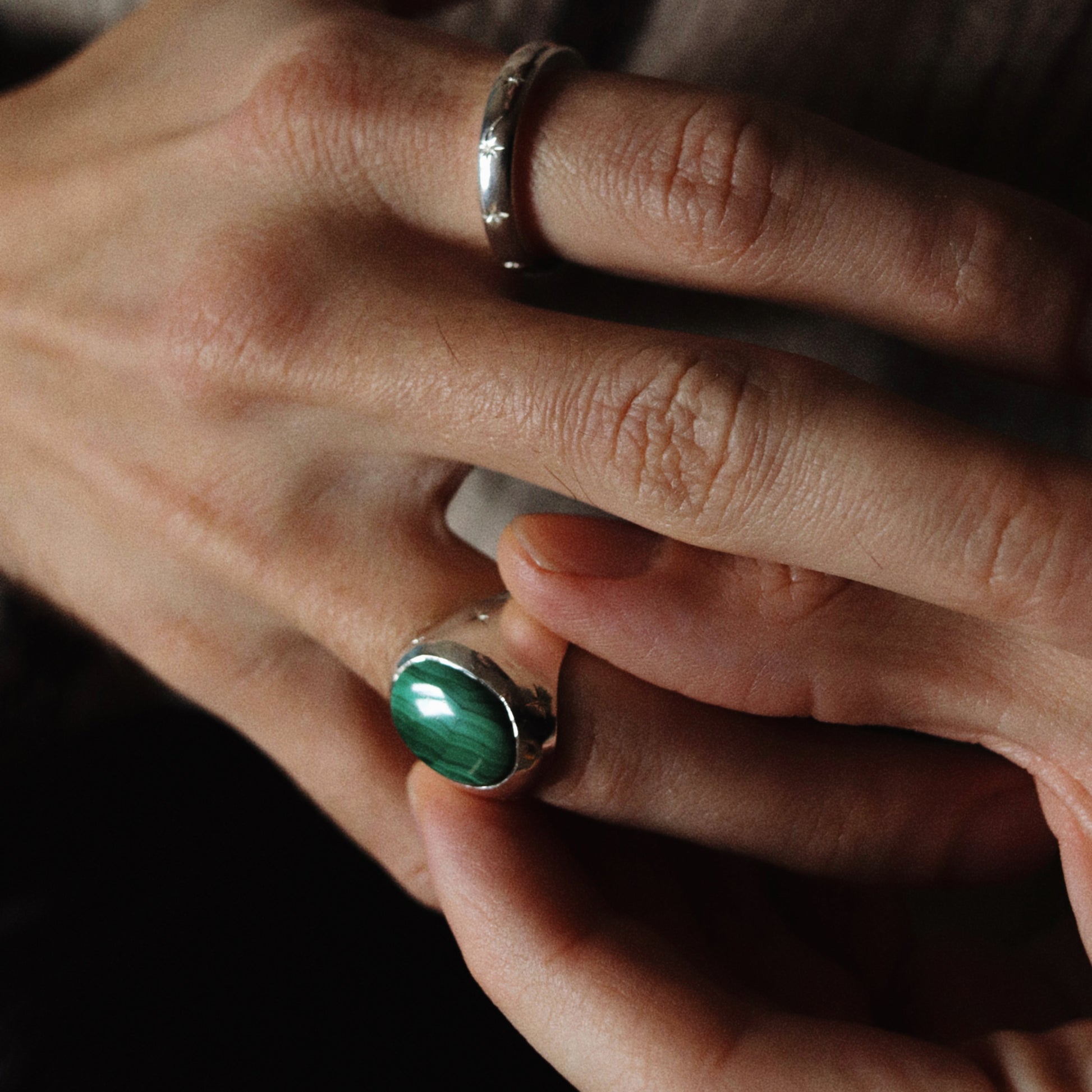 A close-up of a person's hands wearing two rings: a simple silver band on one finger and a silver ring with a large oval green stone on the other finger, set against a dark background and a textured beige sweater.
